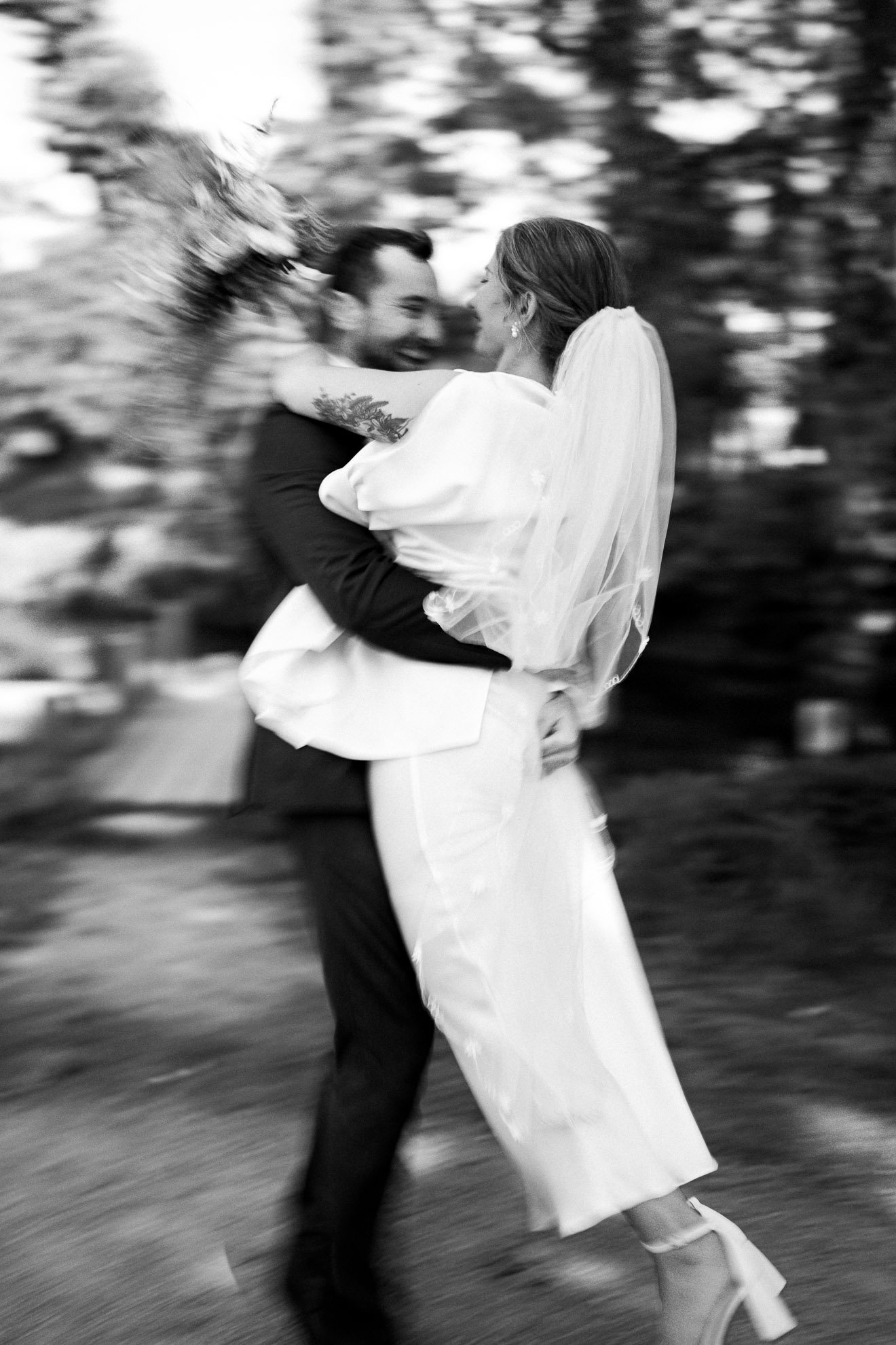 A black and white photo of a couple dancing outside, with the man lifting the woman. The woman is wearing a wedding dress and the man is in a dark suit. They are smiling and close to each other, with blurred background of trees and bushes.