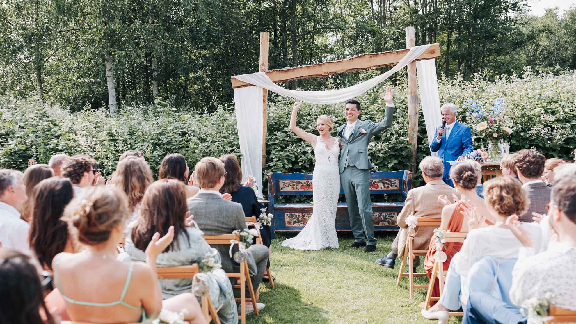 A wedding ceremony outdoors with a bride and groom standing under a wooden arch, smiling and raising their hands in victory, surrounded by seated guests clapping and celebrating, with a officiant speaking nearby, all set in a garden with lush greener