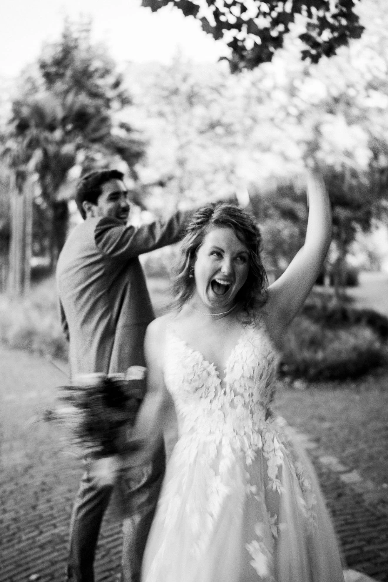 Black and white photo of a woman in a wedding dress laughing and raising her arm, with a man in the background, outdoors near trees and brick pavement.