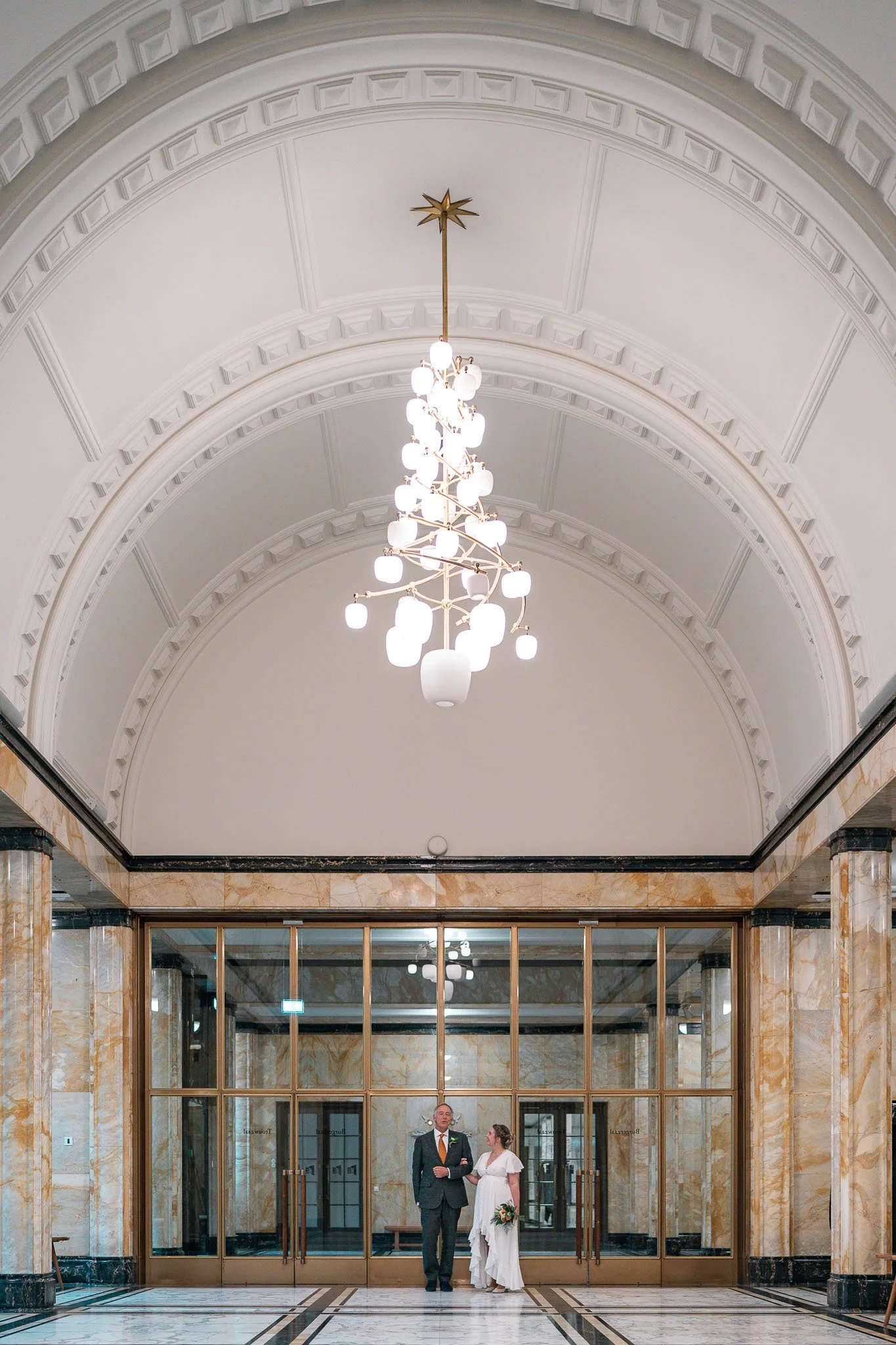 Twee mensen, een man in een pak en een vrouw in een witte jurk, staan arm in arm in een grote, elegante hal met marmeren muren en een hoge witte plafond, verlicht door een grote, moderne chandelier.