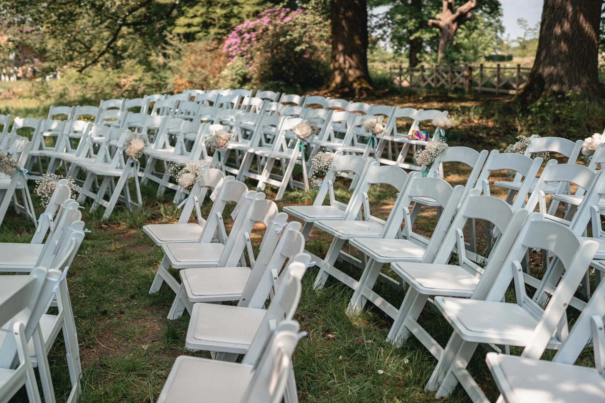 White chairs decorated with floral arrangements set up outdoors for a wedding ceremony, with trees and a wooden fence in the background.