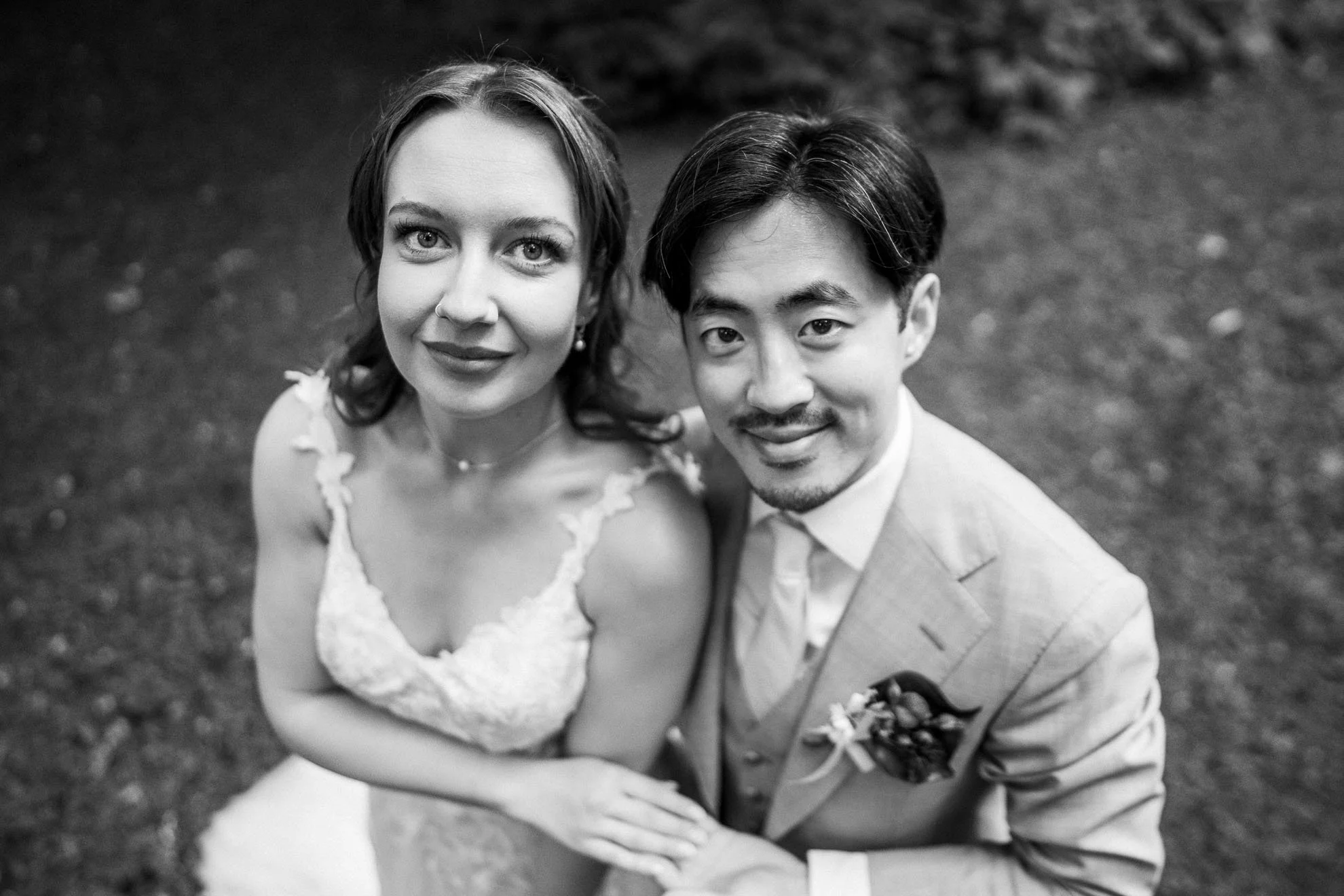 Black and white photograph of a smiling woman and man dressed in wedding attire, looking up at the camera, outdoors on grass.