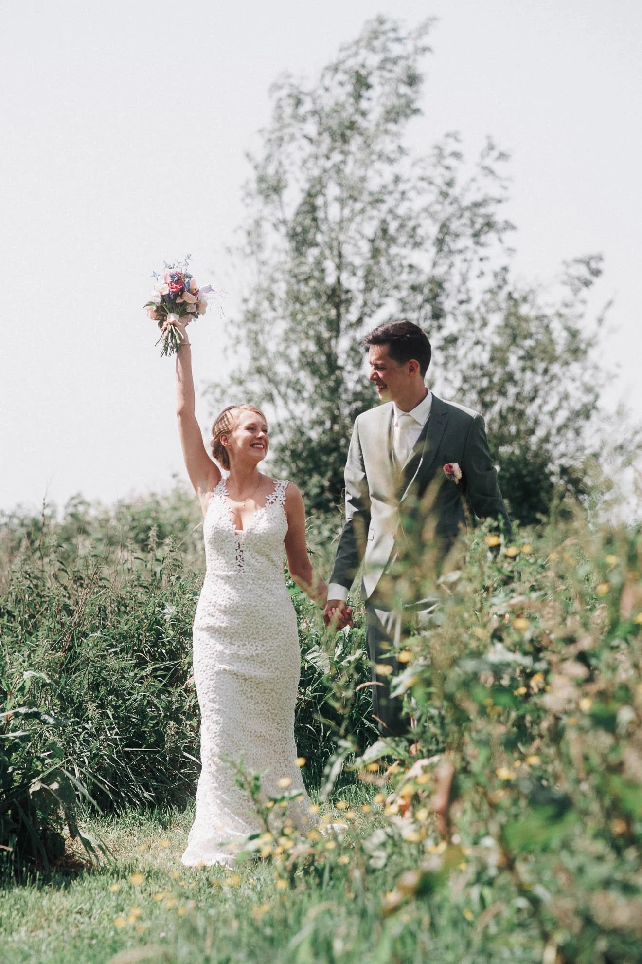 A bride and groom holding hands in a garden, with the bride raising a bouquet in the air and smiling, while the groom looks at her and smiles. The bride is wearing a white lace dress, and the groom is in a gray suit.
