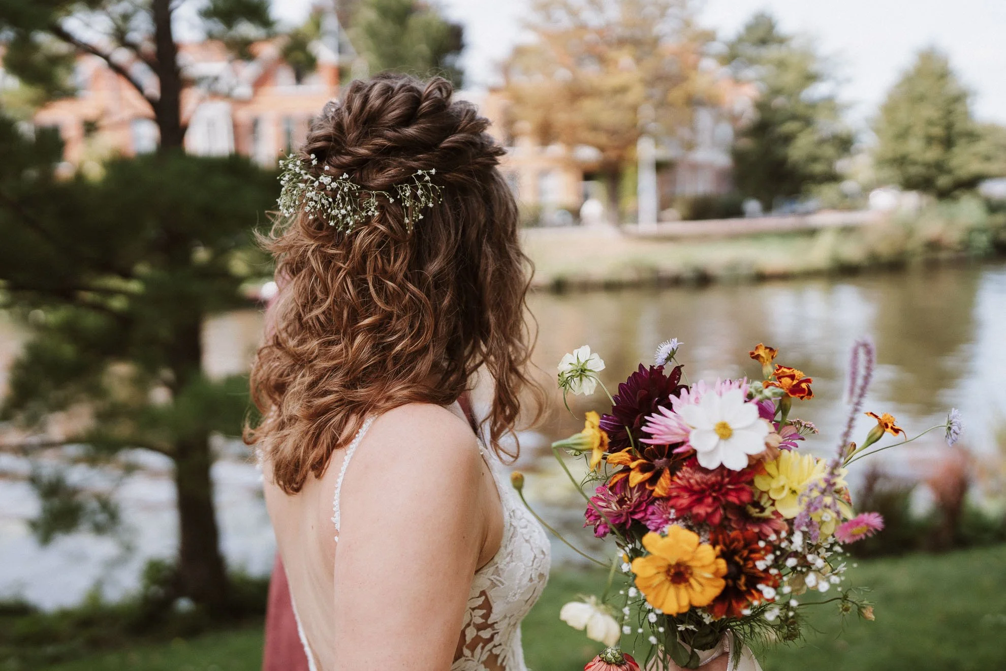 A woman with curly brown hair, decorated with a floral headpiece, holding a colorful bouquet of flowers outdoors near a river with trees and houses in the background.
