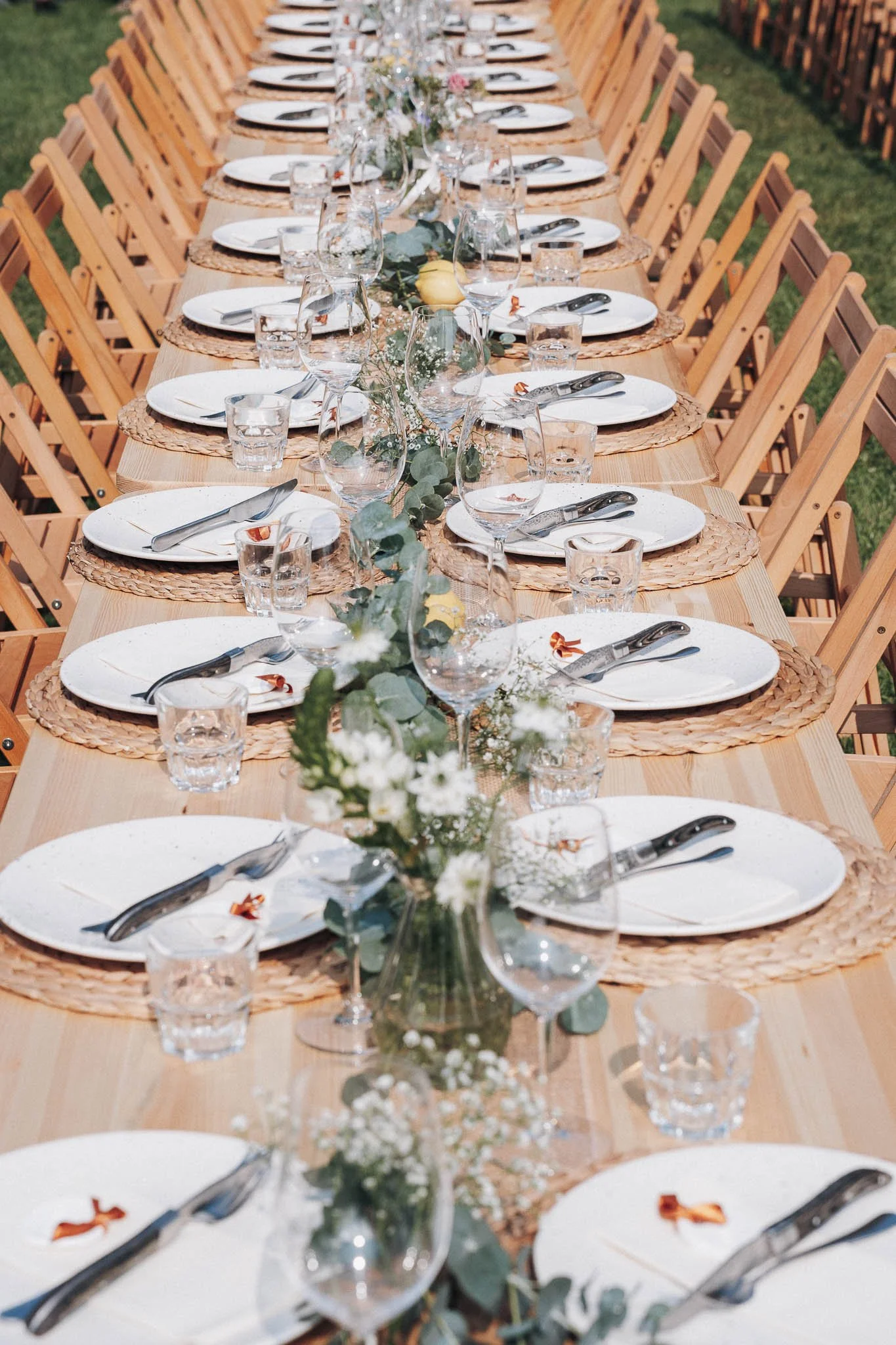 Een lange houten tafel klaar voor een buitenevenement met witte borden, glazen, bestek, en bloemen in vazen als decoratie.