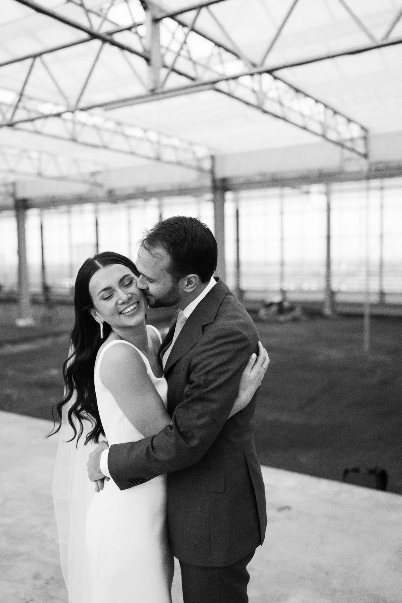 A black and white photo of a couple embracing in a large, industrial-style indoor space with metal beams and large windows.