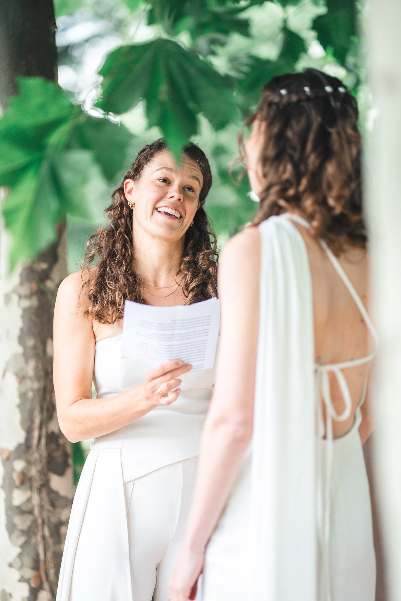 Two women with curly hair, one holding a paper, talking outdoors among large green leaves.