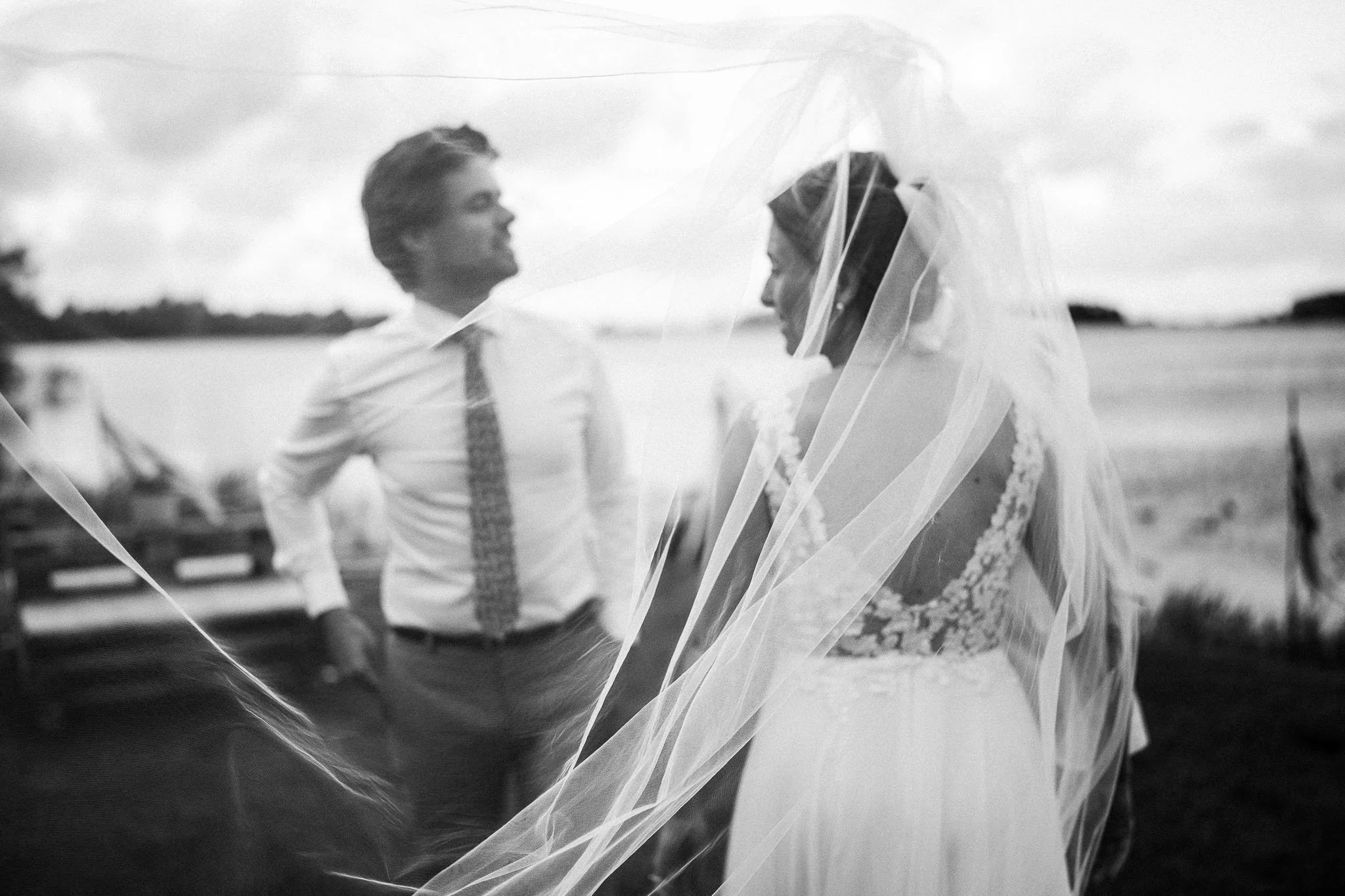 Een bruid en een bruidegom staan samen buiten, de bruid heeft een sluier over haar hoofd en het uitzicht op de achtergrond is een waterpartij en een weide.