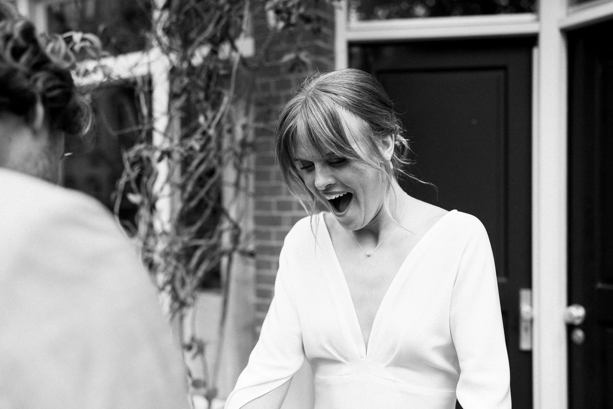 Black and white photo of a woman with short hair smiling and looking down, wearing a white top, outdoors with brick wall and plants in the background.