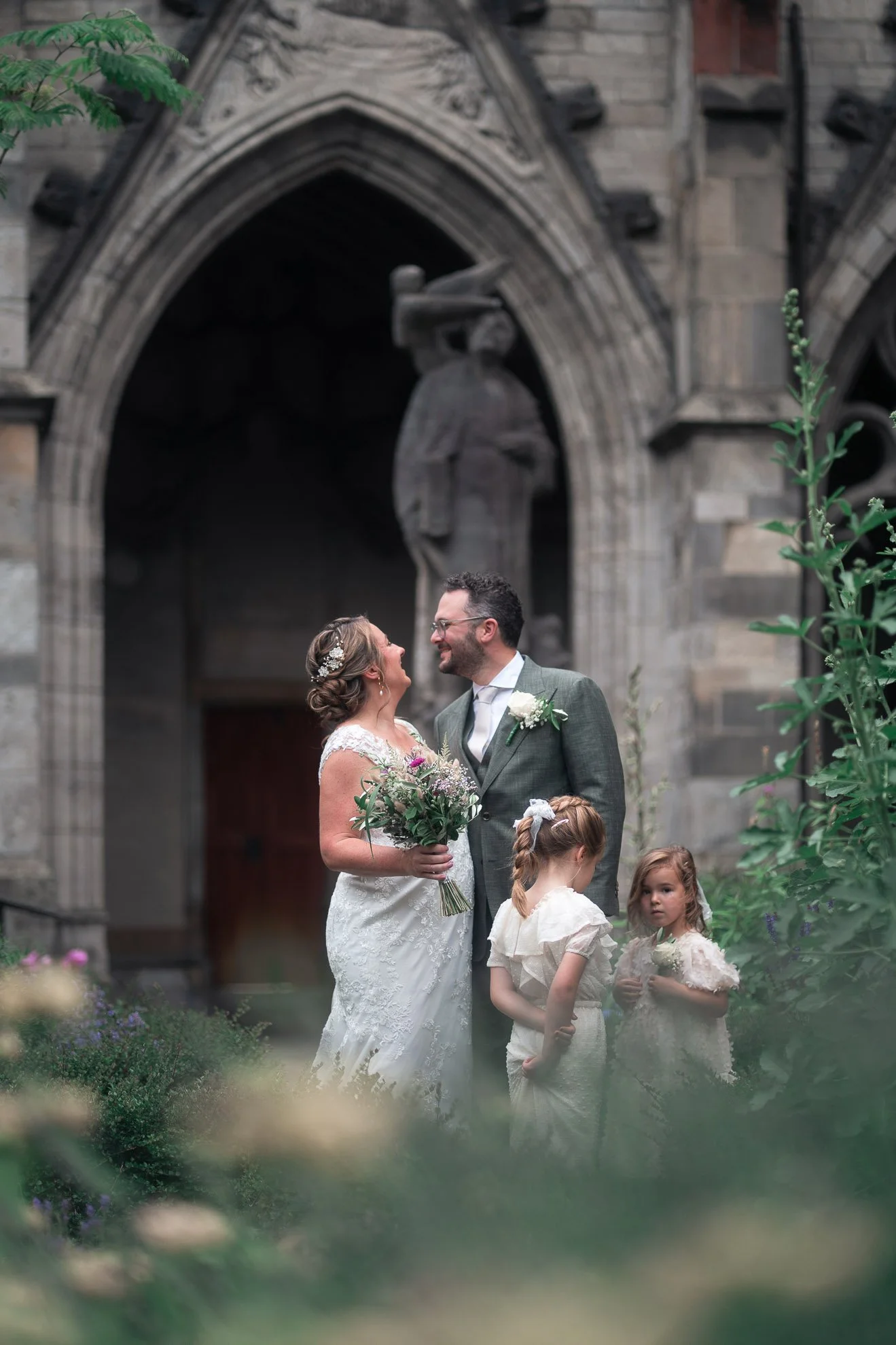 A bride and groom standing outside in front of a stone church, smiling and looking at each other, with two young girls standing nearby, all dressed in formal clothing for a wedding ceremony.