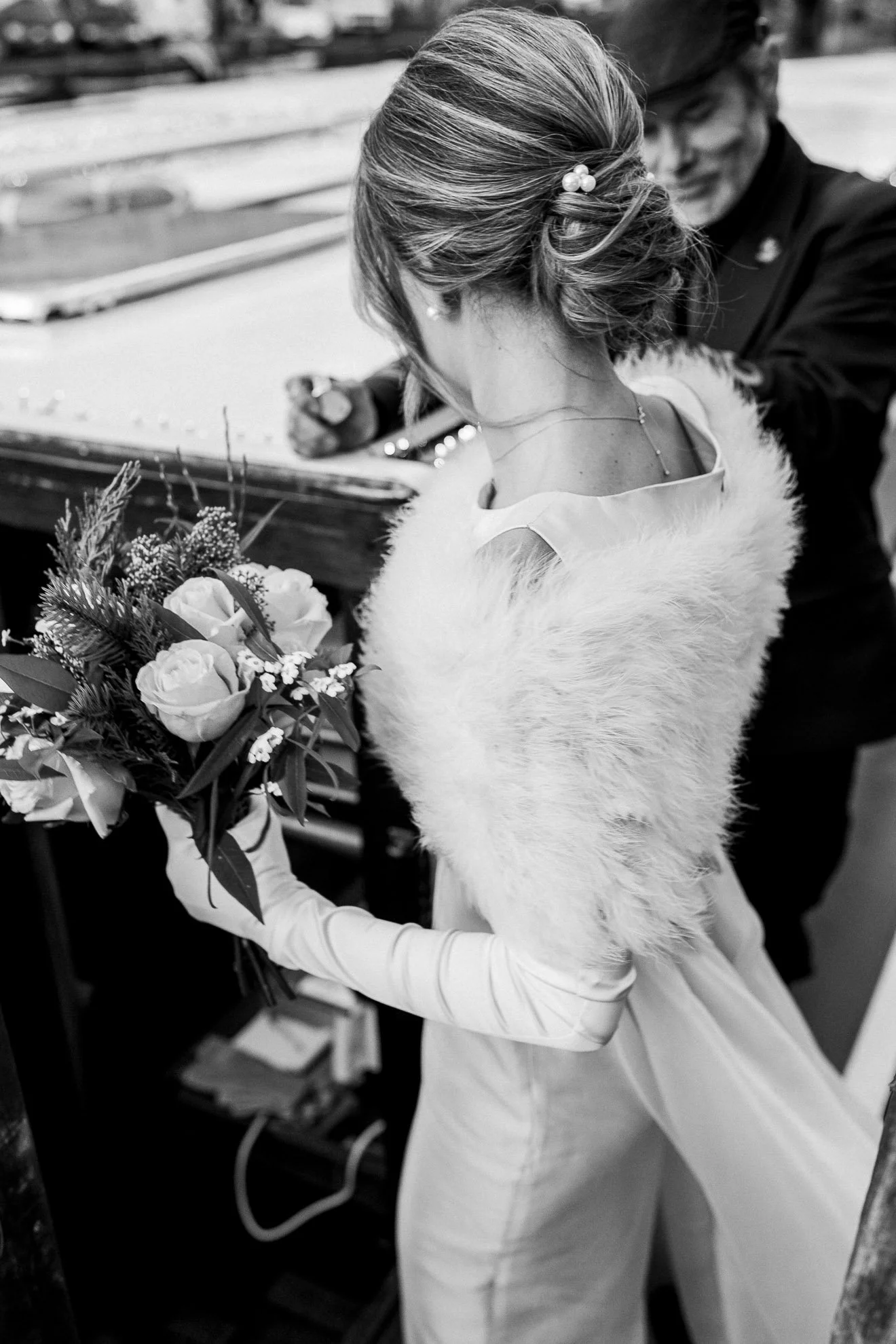 A bride in a wedding dress and veil is holding a bouquet of flowers, standing next to a woman in a black outfit who is signing or writing on a surface.