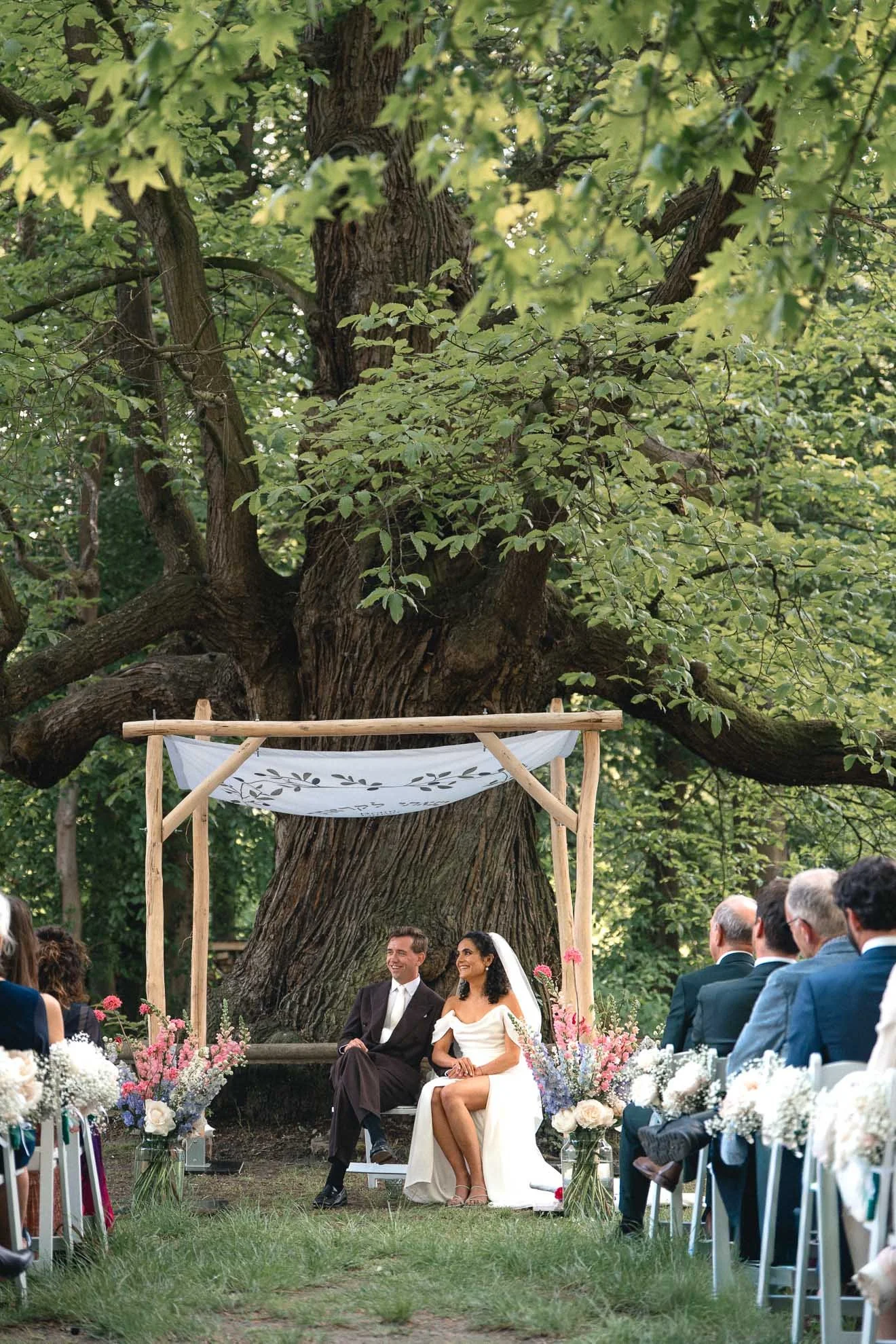 A wedding ceremony outdoors under a large tree with a wooden frame and white fabric. The bride and groom are seated in front of a crowd of wedding guests, surrounded by colorful flowers in vases. The bride is wearing a white dress and veil, and the g
