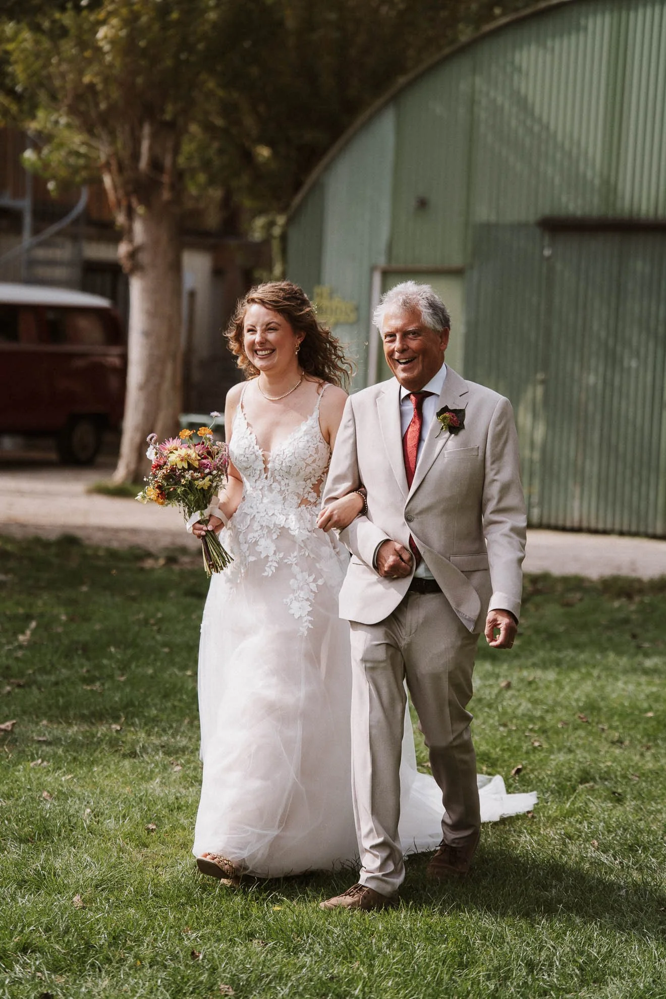 A bride in a white lace wedding dress holding a bouquet walking with an older man in a light gray suit, outdoors on grass with trees and a green building in the background.