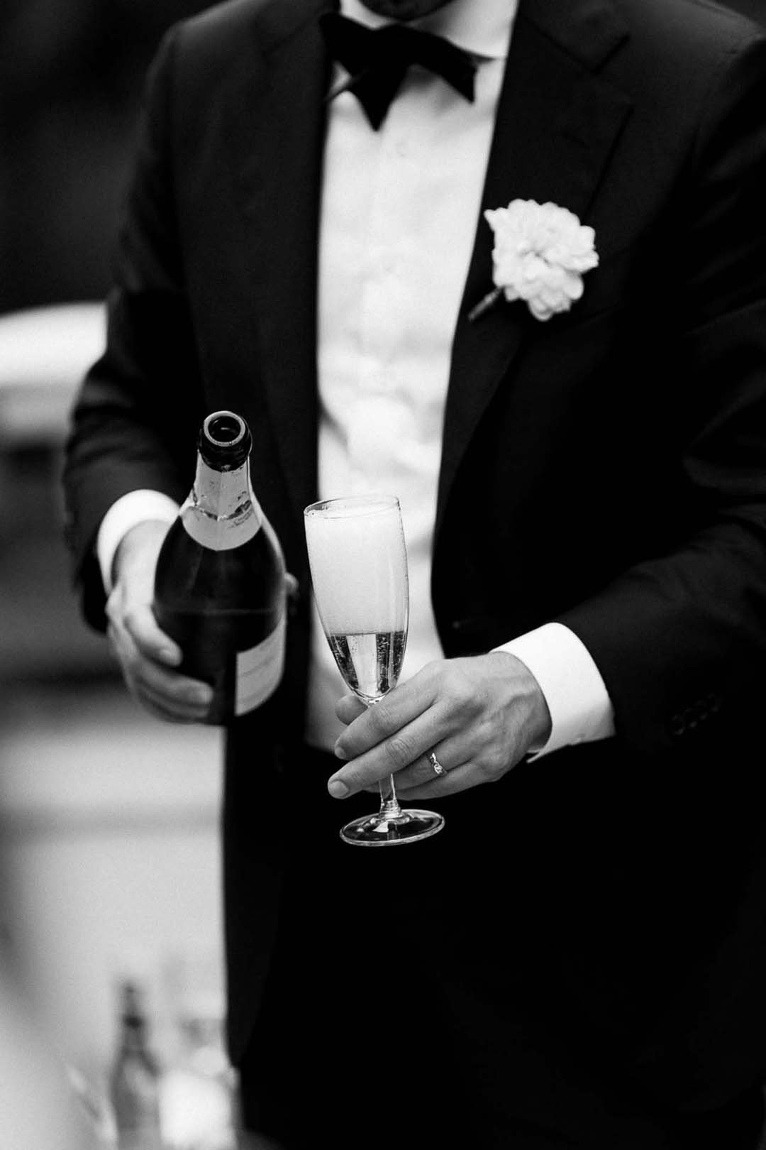 Man in smoking met witte blouse, zwarte blazer en bloemenlint, schenkt champagne in een glas.
