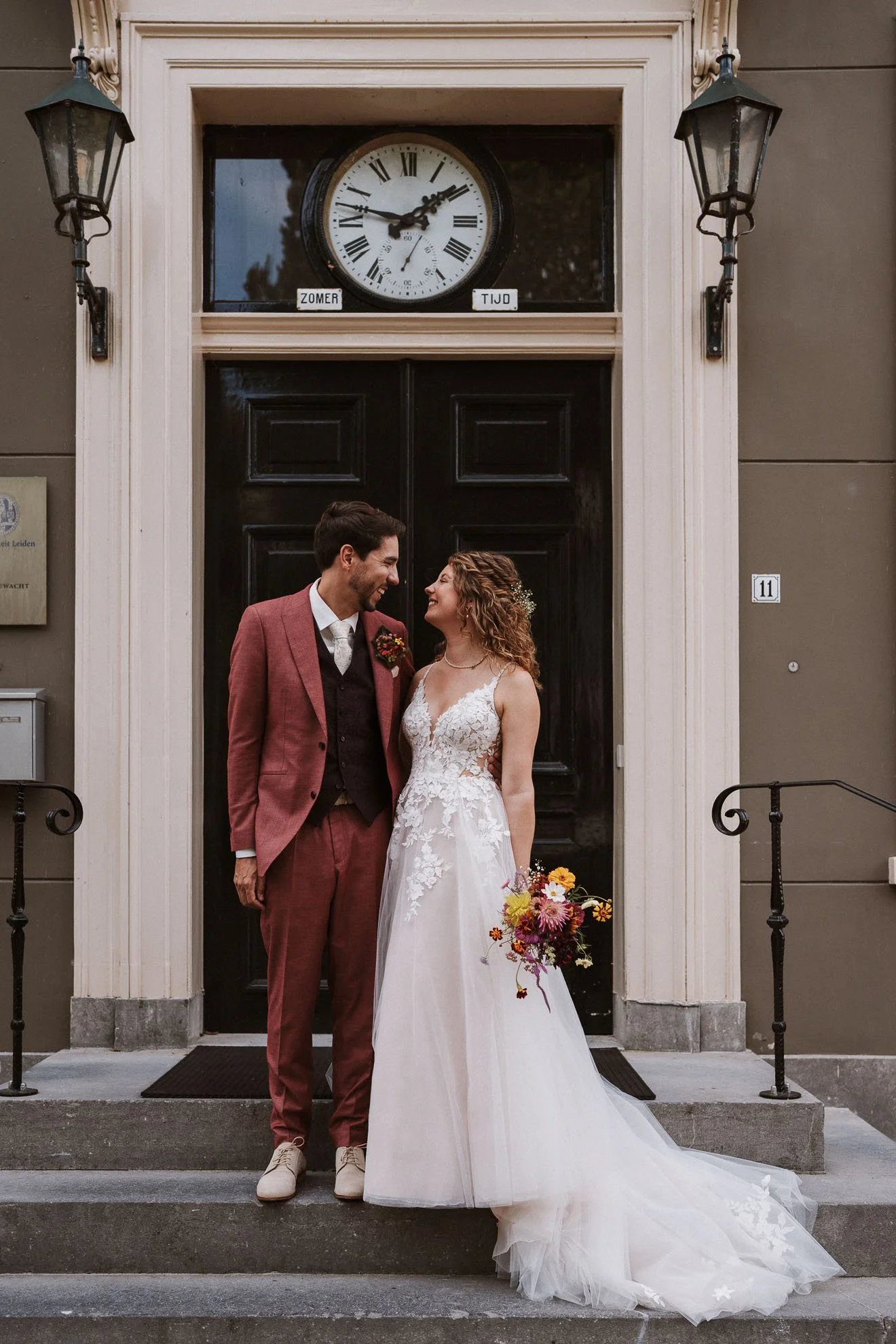 A bride and groom stand on the steps in front of a black door, smiling and looking at each other. The groom is wearing a mauve suit, white shirt, and brown shoes, and is holding a bouquet. The bride is wearing a white lace wedding gown and holding a 