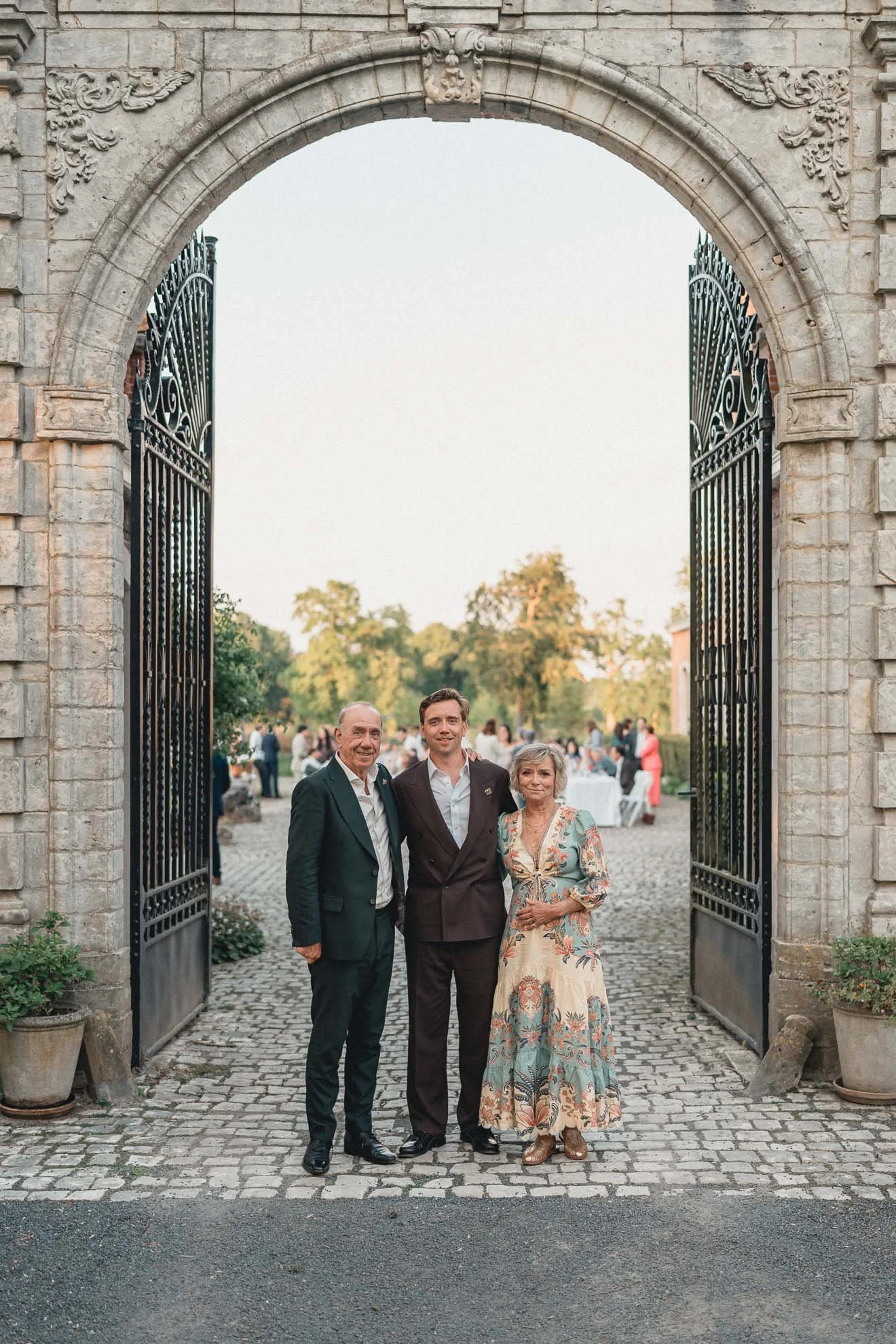 Three people standing under a large stone archway with open iron gates, at an outdoor event with tables and people in the background during sunset.