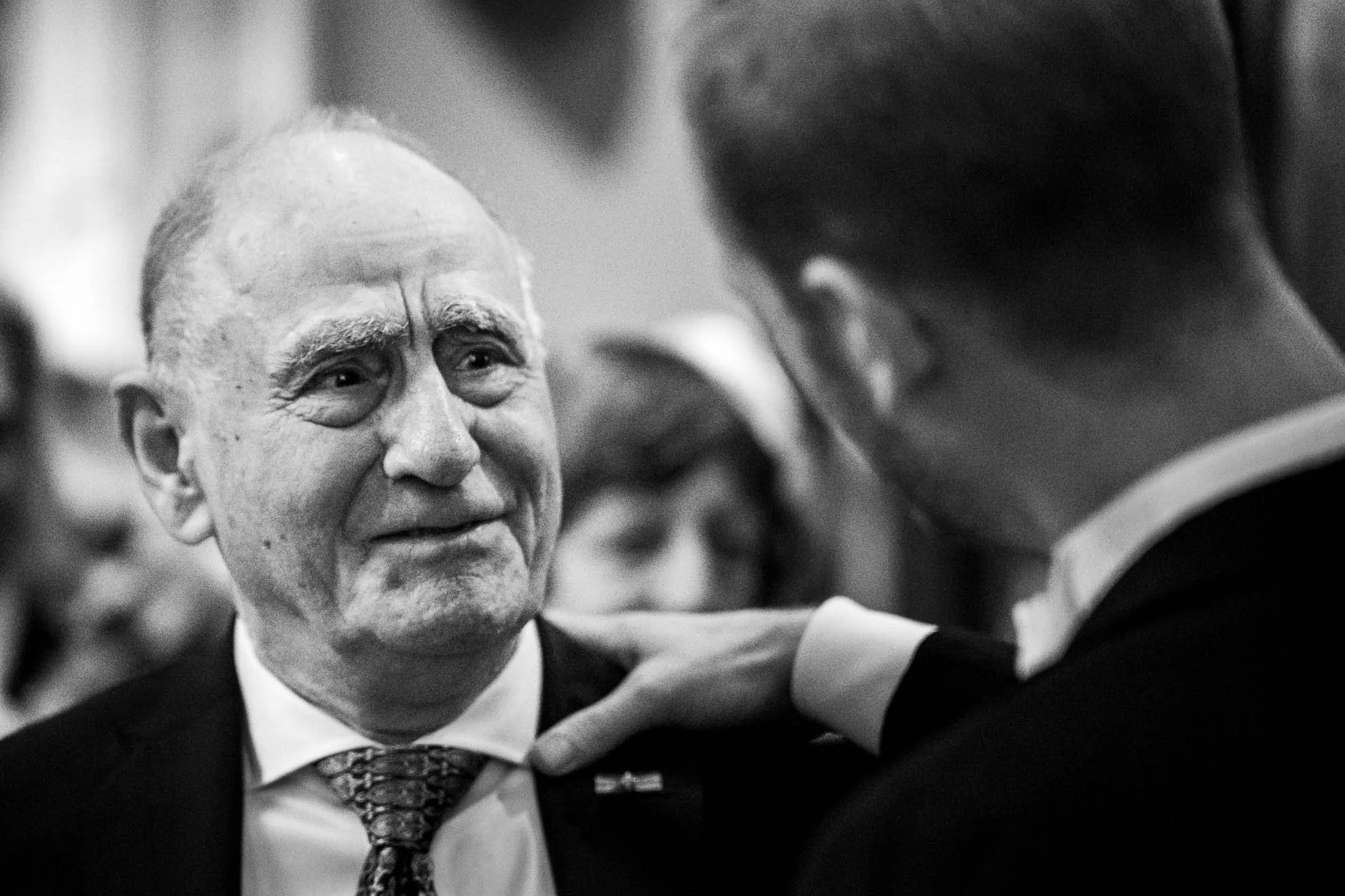 An elderly man with a bald head and gray beard is smiling while looking at a young man in a suit who is saluting him. The photo appears to be taken in a formal setting, possibly a ceremony or event.