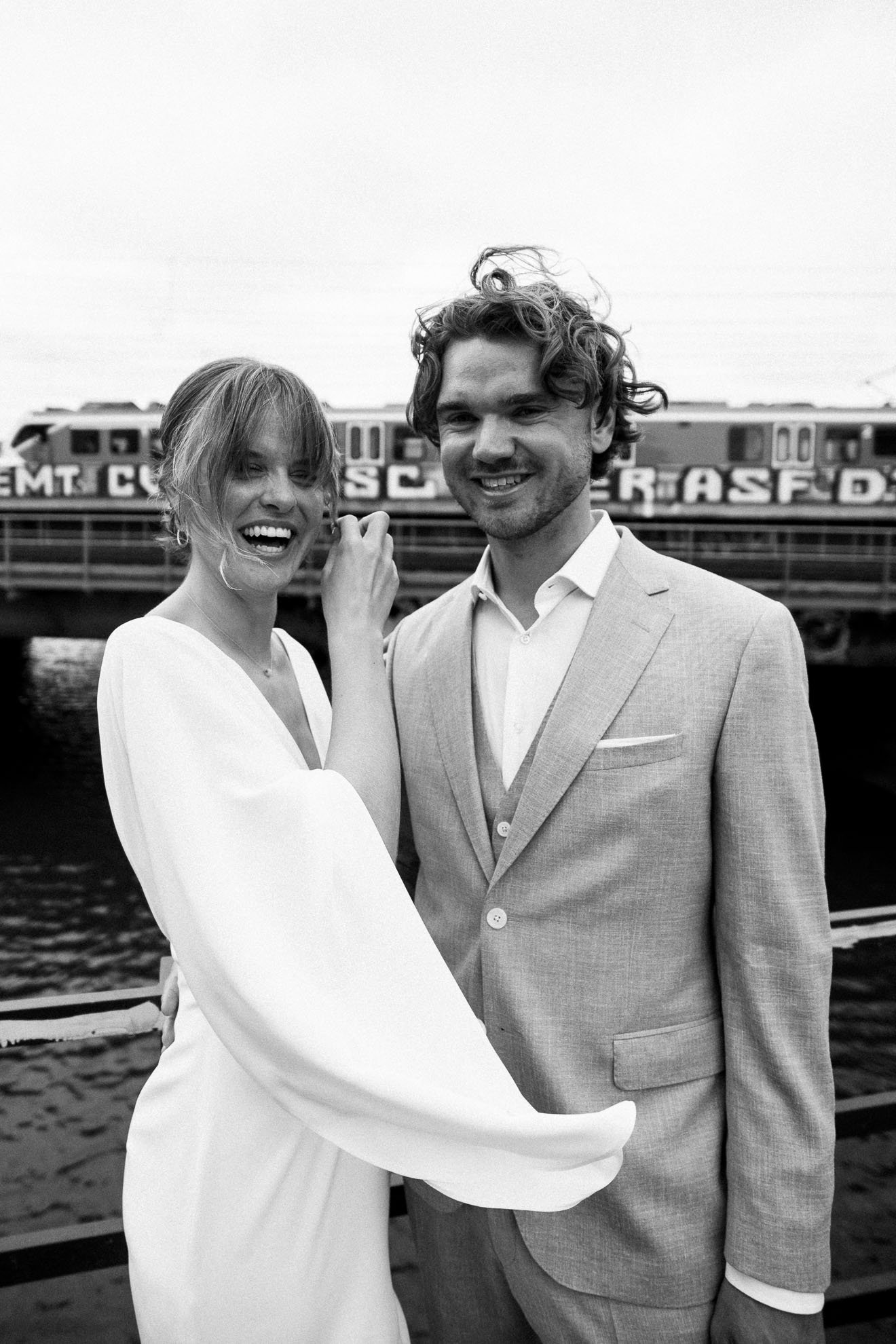 A black and white photograph of a smiling man and woman standing outdoors in front of a train. The woman is wearing a white dress, and the man is wearing a light-colored suit.