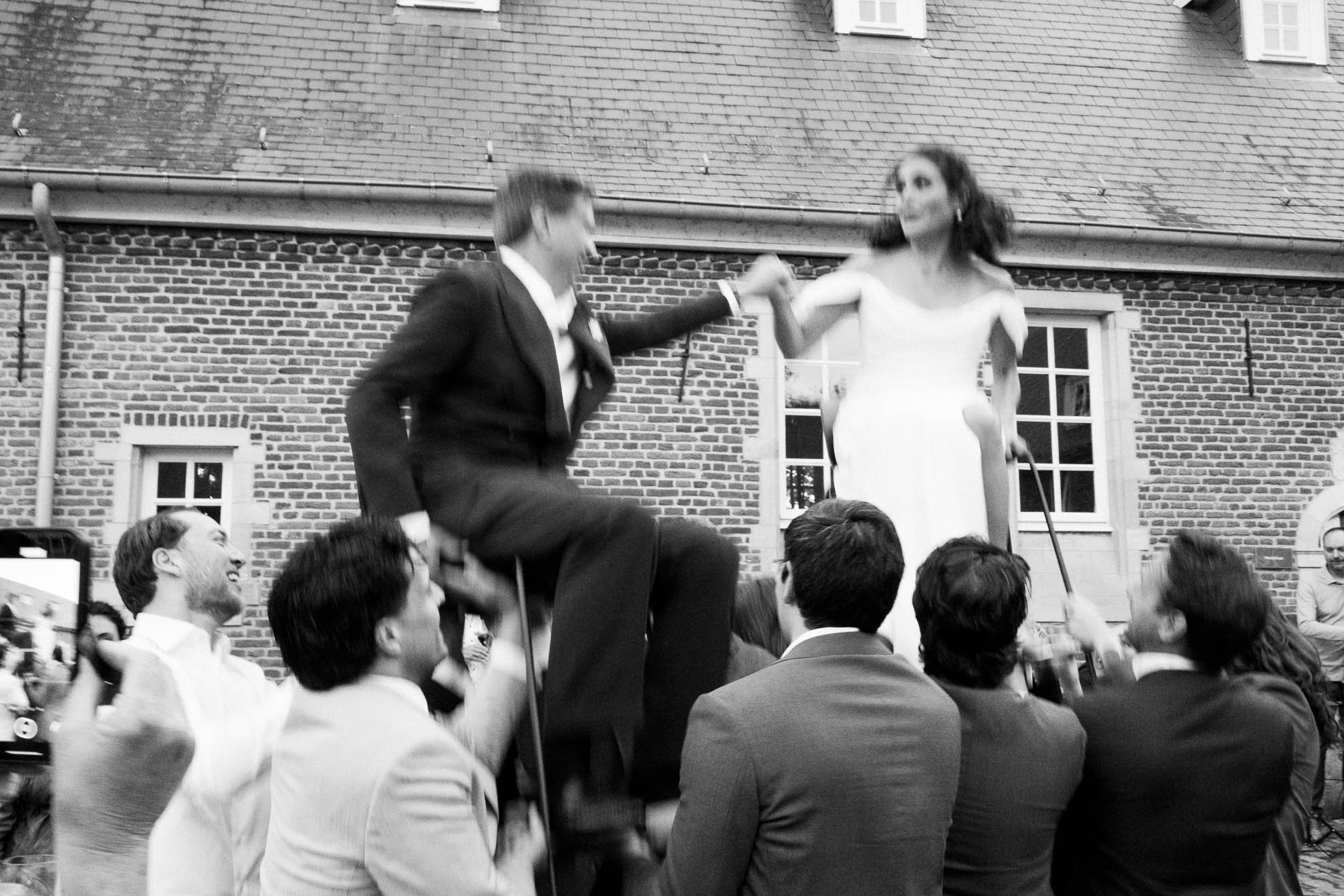 A black and white photograph of a crowd at an event outdoors in front of a brick building with windows. Two people are prominent in the center, a man in a suit sitting on a person's shoulders holding a woman's hand. The woman is wearing a white dress