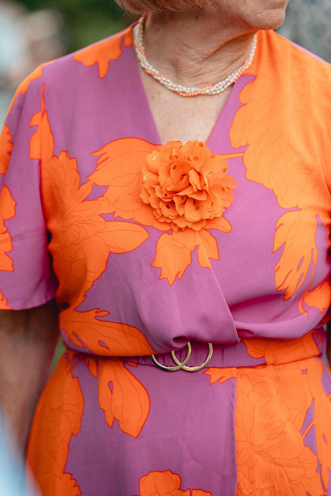Close-up of a woman wearing a bright pink and orange floral dress with a large orange fabric flower pin on the chest, a pearl necklace, and a gold belt with a linked double-ring buckle.