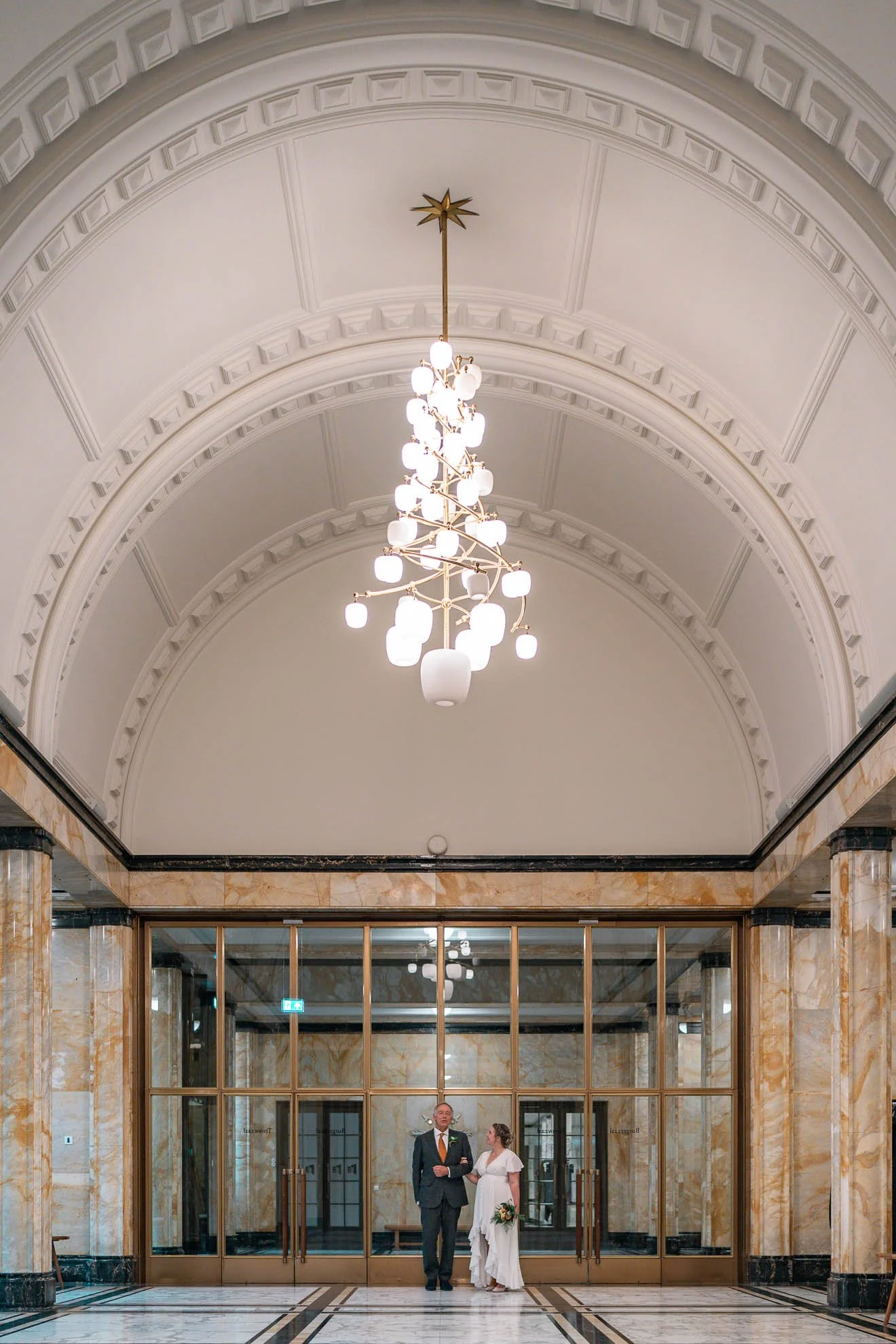 A man and a woman, dressed in formal wedding attire, stand under a large chandelier in an elegant, marble-lined building with arched ceilings and glass doors.