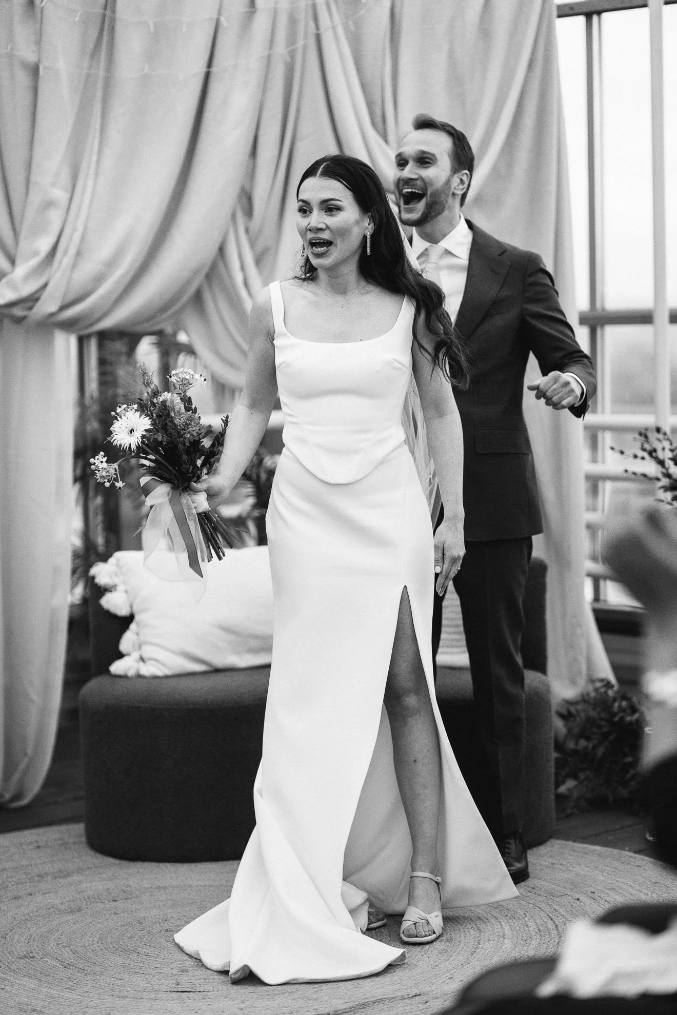 Black and white photo of a bride and groom during a wedding ceremony. The bride is wearing a white wedding dress with a slit, holding a bouquet, and standing in front of a decorated arch. The groom is behind her, wearing a suit, smiling, and looking 
