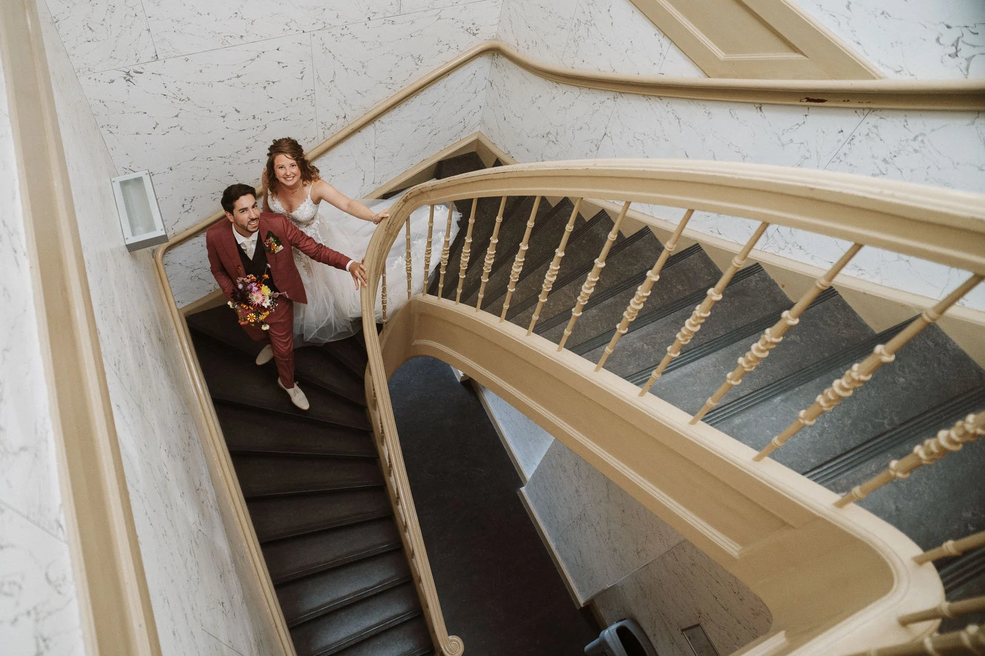 A bride and groom standing on a staircase looking up at a camera. The bride is wearing a white wedding dress, and the groom is dressed in a suit with a bouquet of flowers. The staircase has a wooden handrail and the floor is marble.