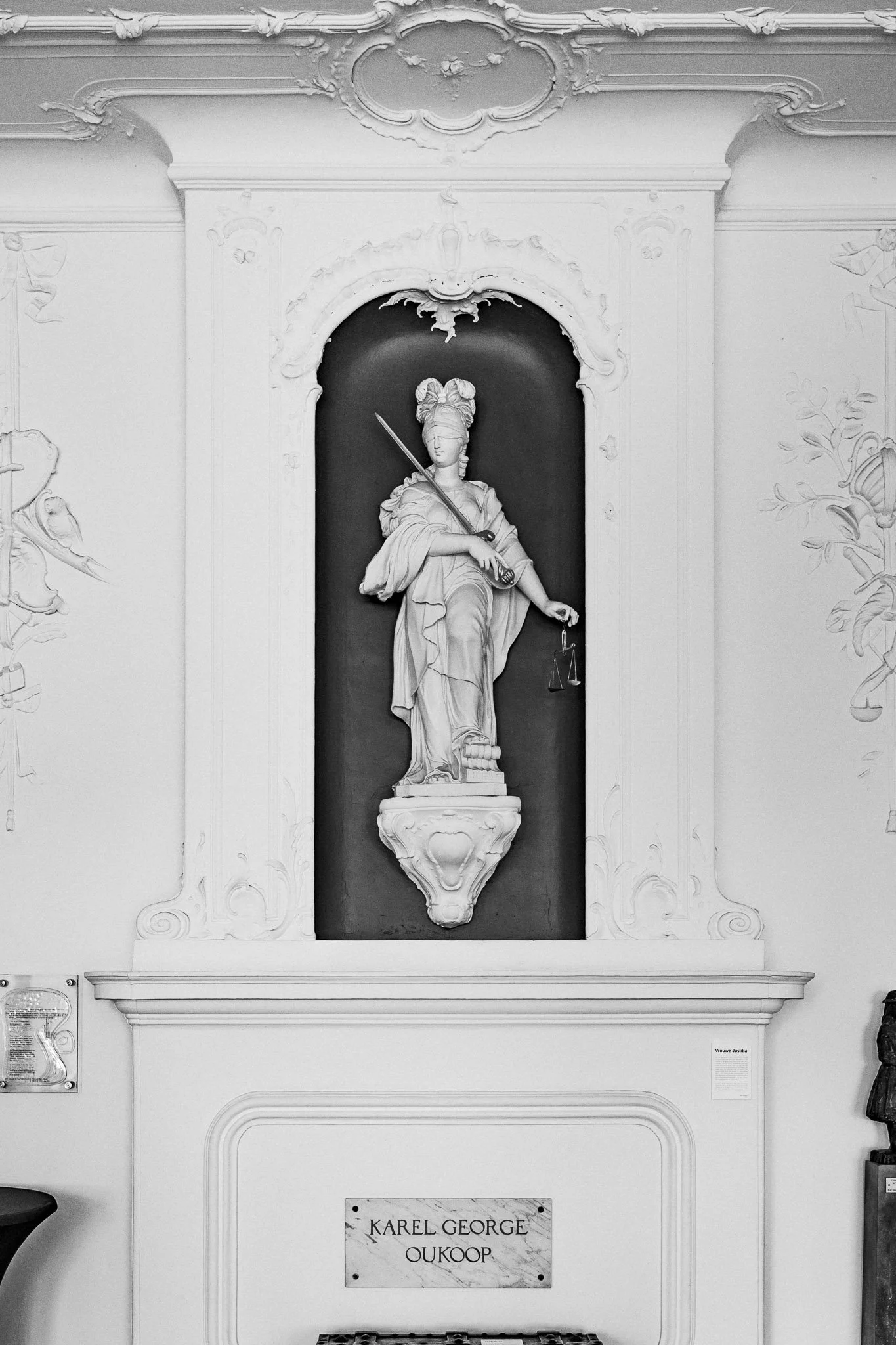 Een zwart-wit afbeelding van een beeldhouwwerk van een vrouwelijke figuur met een zwaard en een weegschaal, geplaatst in een nis met decoratieve elementen eromheen.