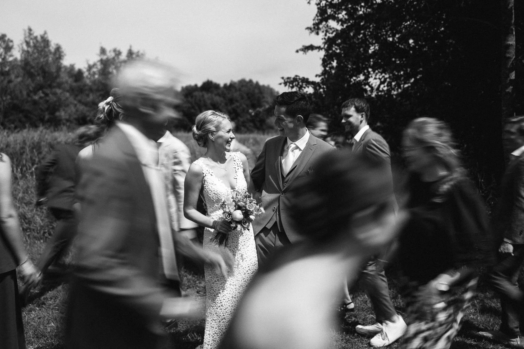 Black and white photo of a wedding ceremony outdoors with people dancing around a bride and groom, celebrating and smiling.