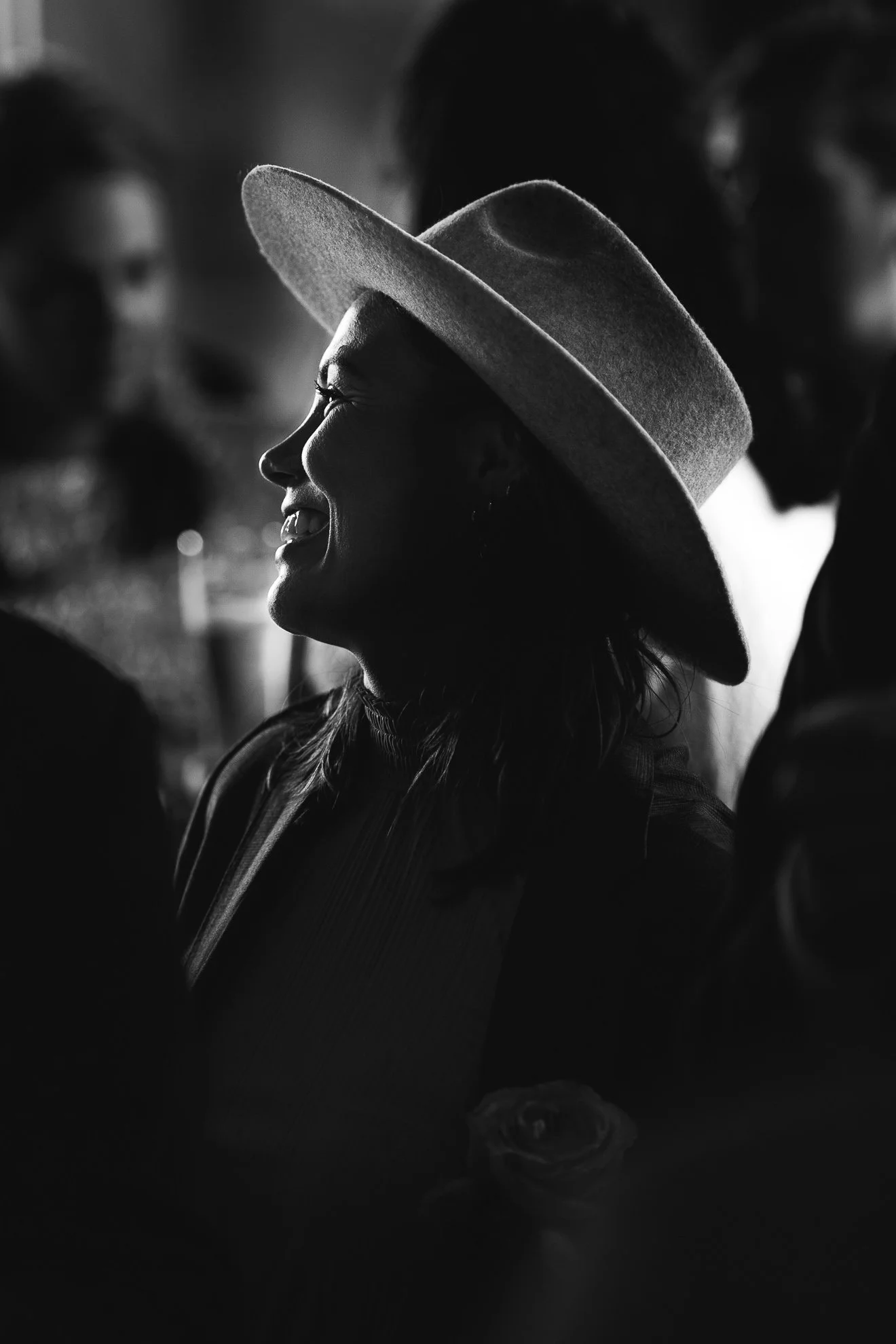 Black and white photo of a woman wearing a wide-brimmed hat, smiling, with dark hair, in a social setting with blurred background.