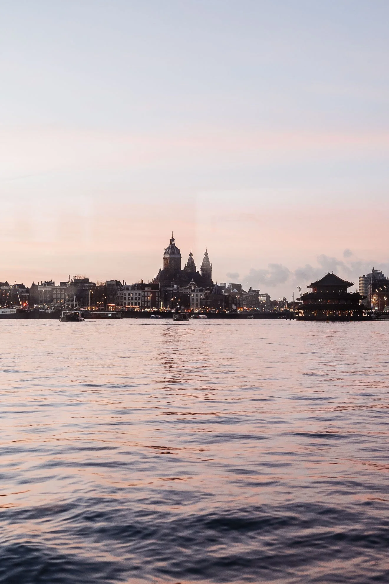 A city skyline across a body of water during sunset, with a historic church and other buildings, and a pagoda-style structure on the right.