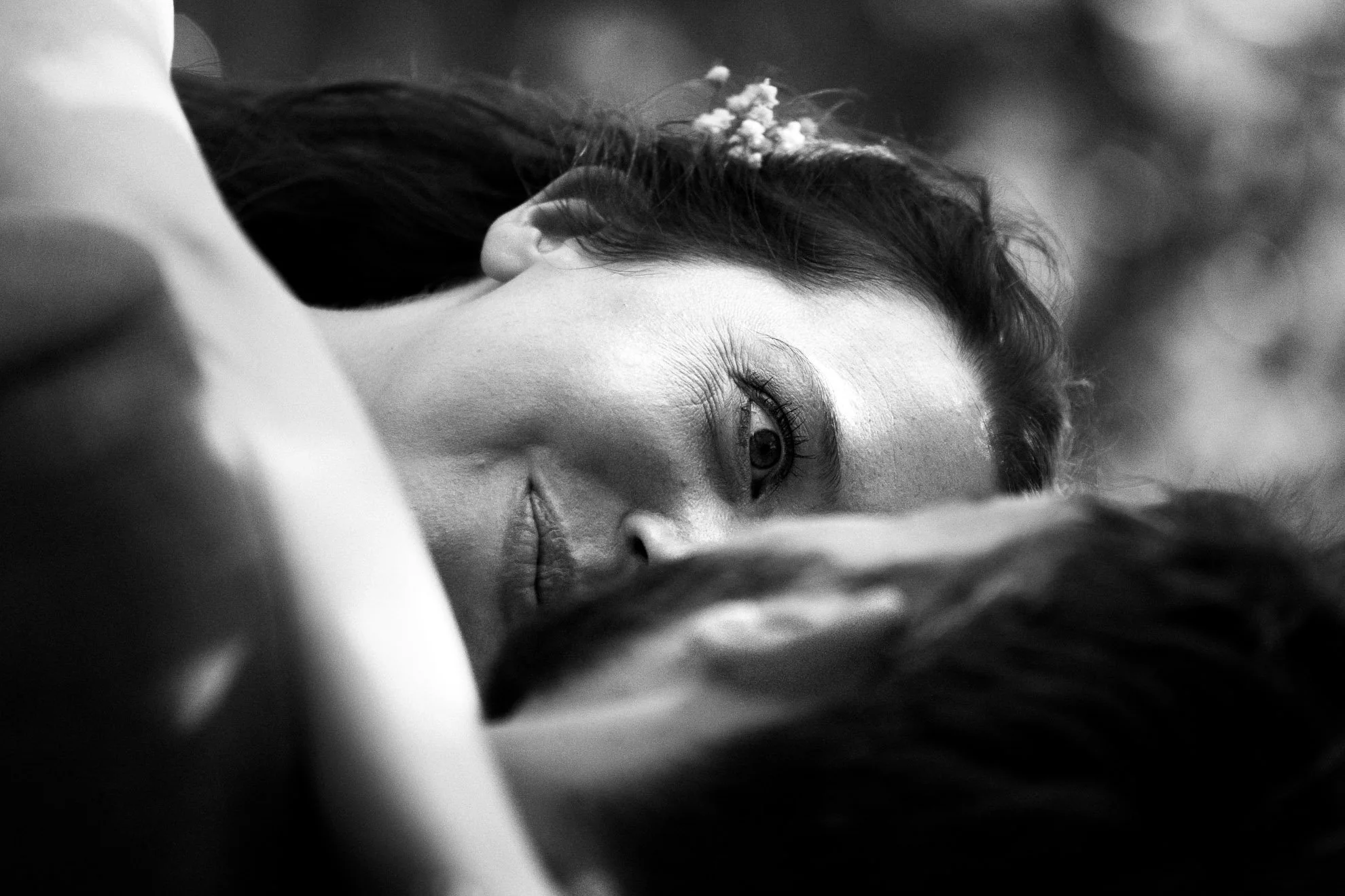 A close-up black and white photo of a woman lying down, partially covered by a surface, with her face turned slightly and eyes looking at the camera. She has dark hair with small flowers in it.
