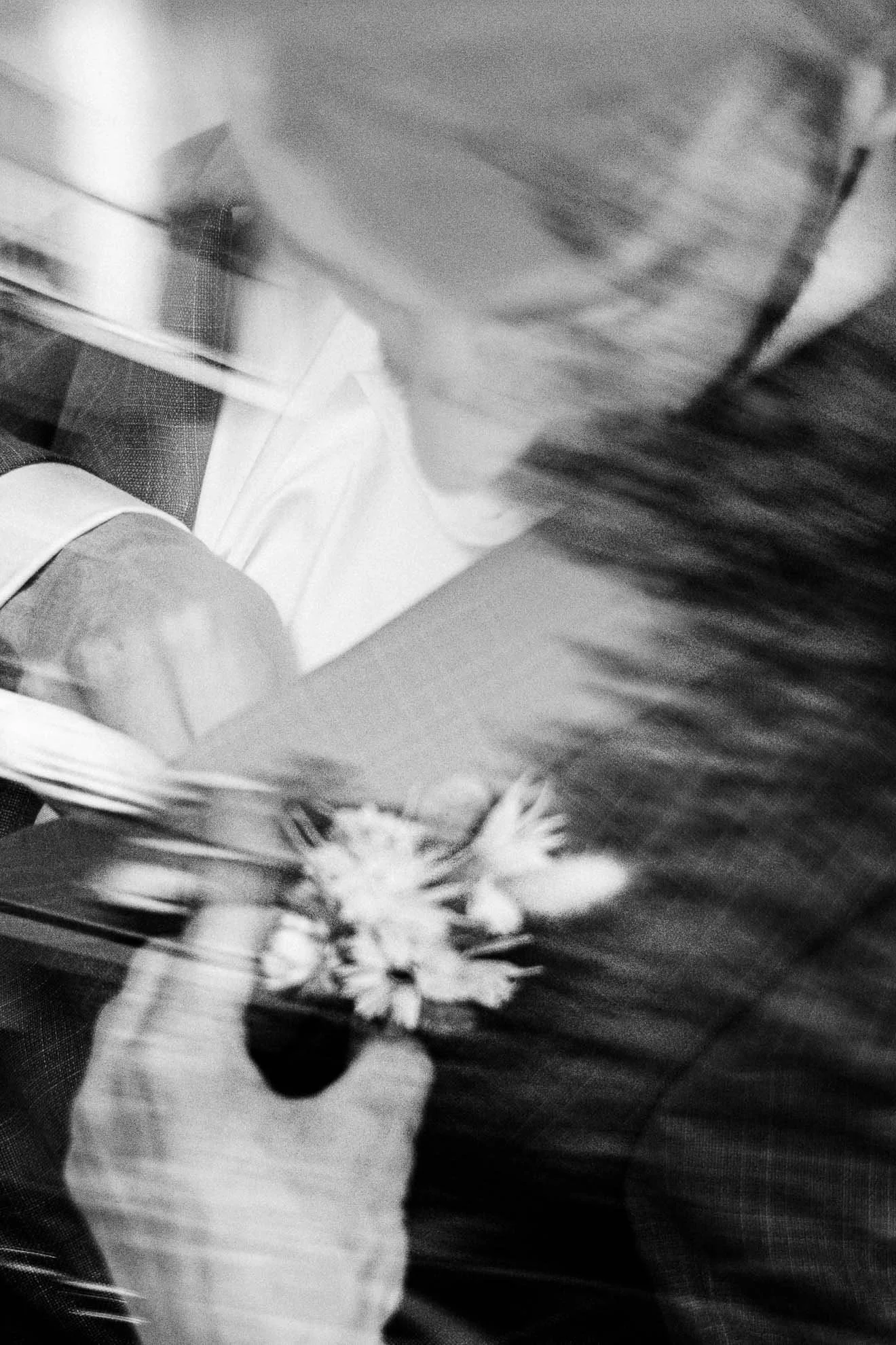 Black and white photo of a person holding a bouquet of flowers. The person's arm and hand are visible, but their face is not. The background includes some patterned flooring or table. The photo has a slightly blurry, grainy quality.