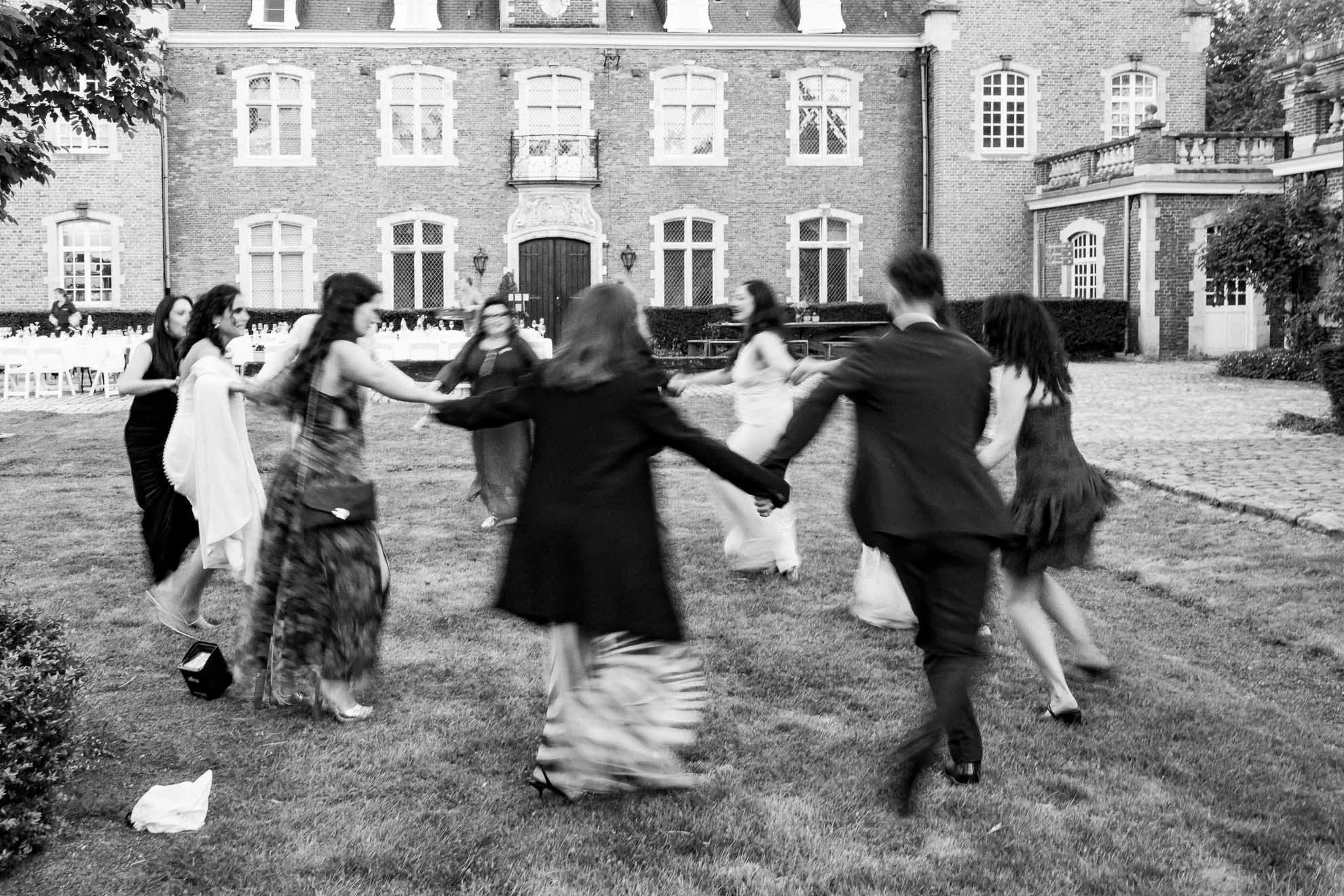 A group of people dancing in a circle on a lawn outside a large brick building.