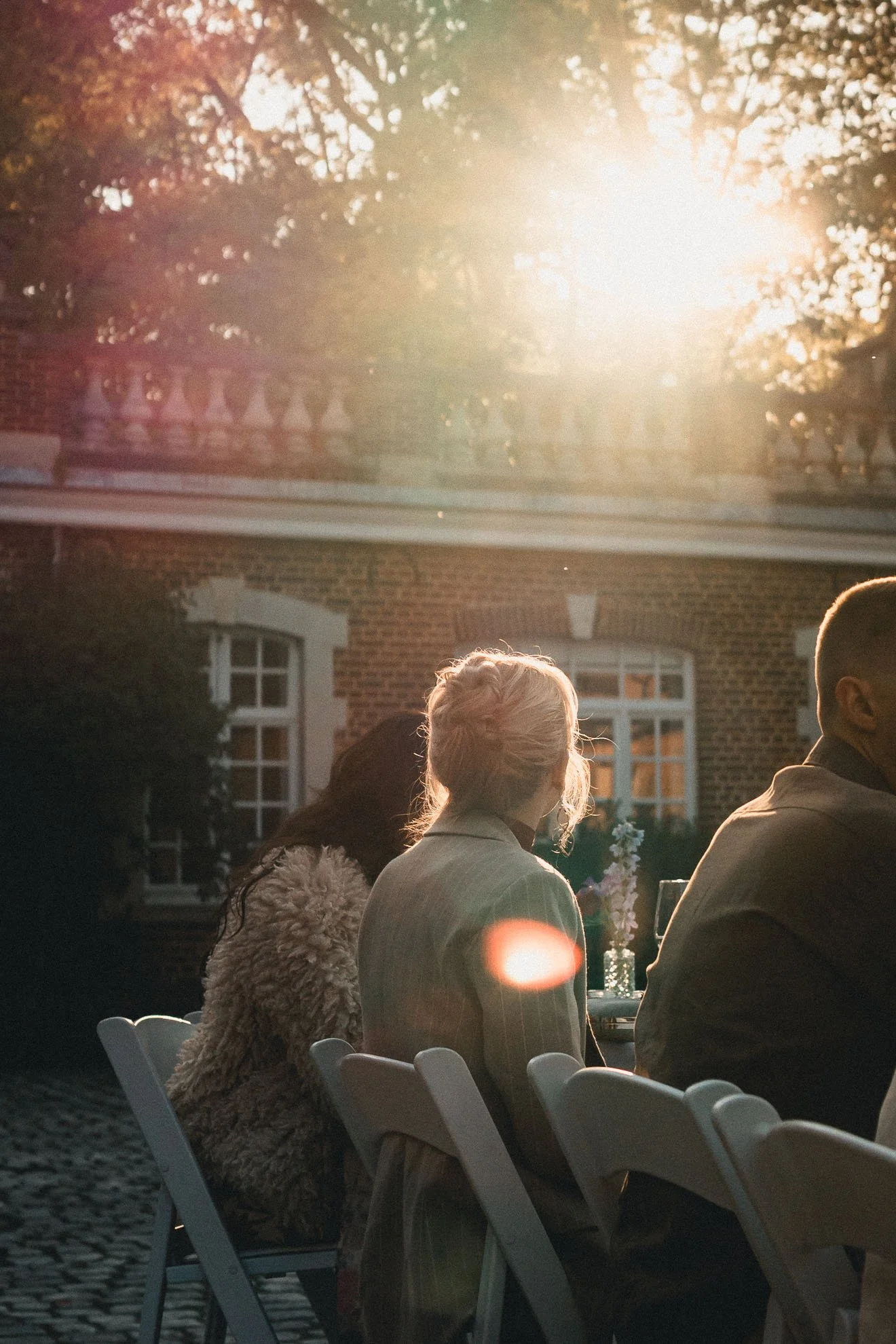 People sitting outdoors at tables during a sunset, with a brick building and trees in the background. The sunlight creates a warm glow and lens flare.