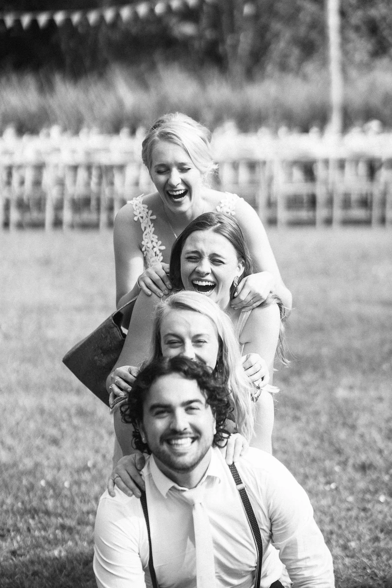 Vijf mensen in een rij die een vriendelijke sprong maken en lachen, buiten op gras, met een rij houten stoelen op de achtergrond, zwart-wit foto.