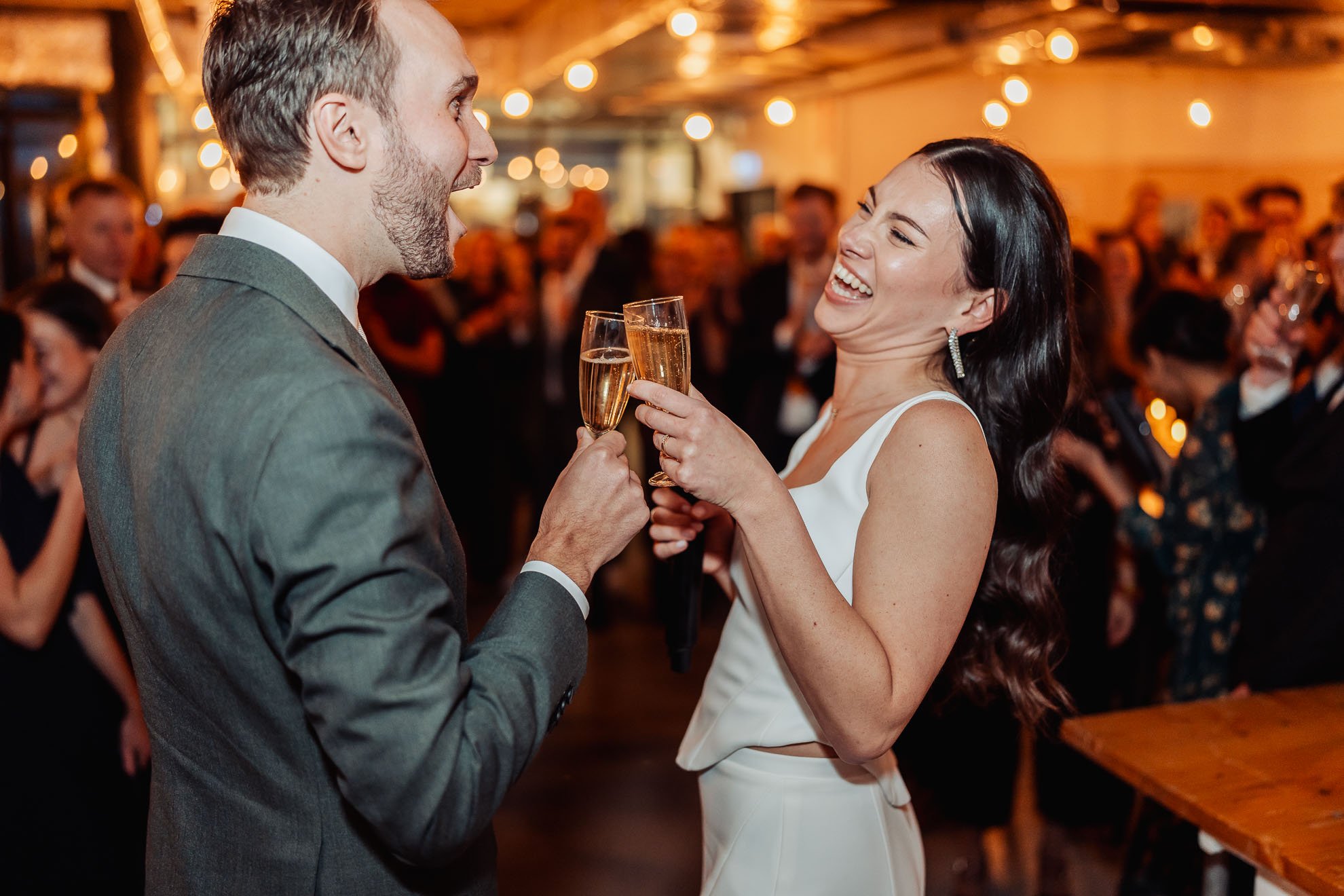 A man and woman at a celebration, smiling and toasting with champagne glasses. They are dressed in formal attire and are surrounded by a crowd in a warmly lit venue.