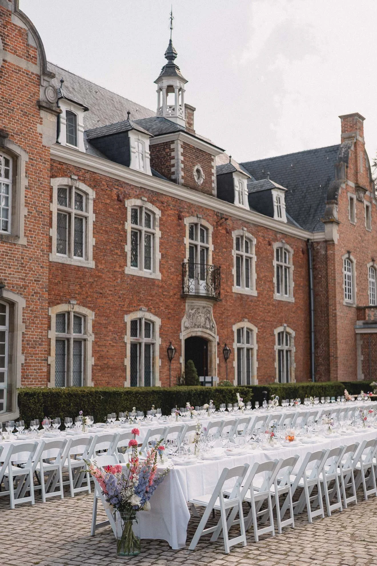 Outdoor wedding reception setup with long white tables and white chairs, decorated with floral arrangements, in front of a historic red brick building with multiple windows and a tower.