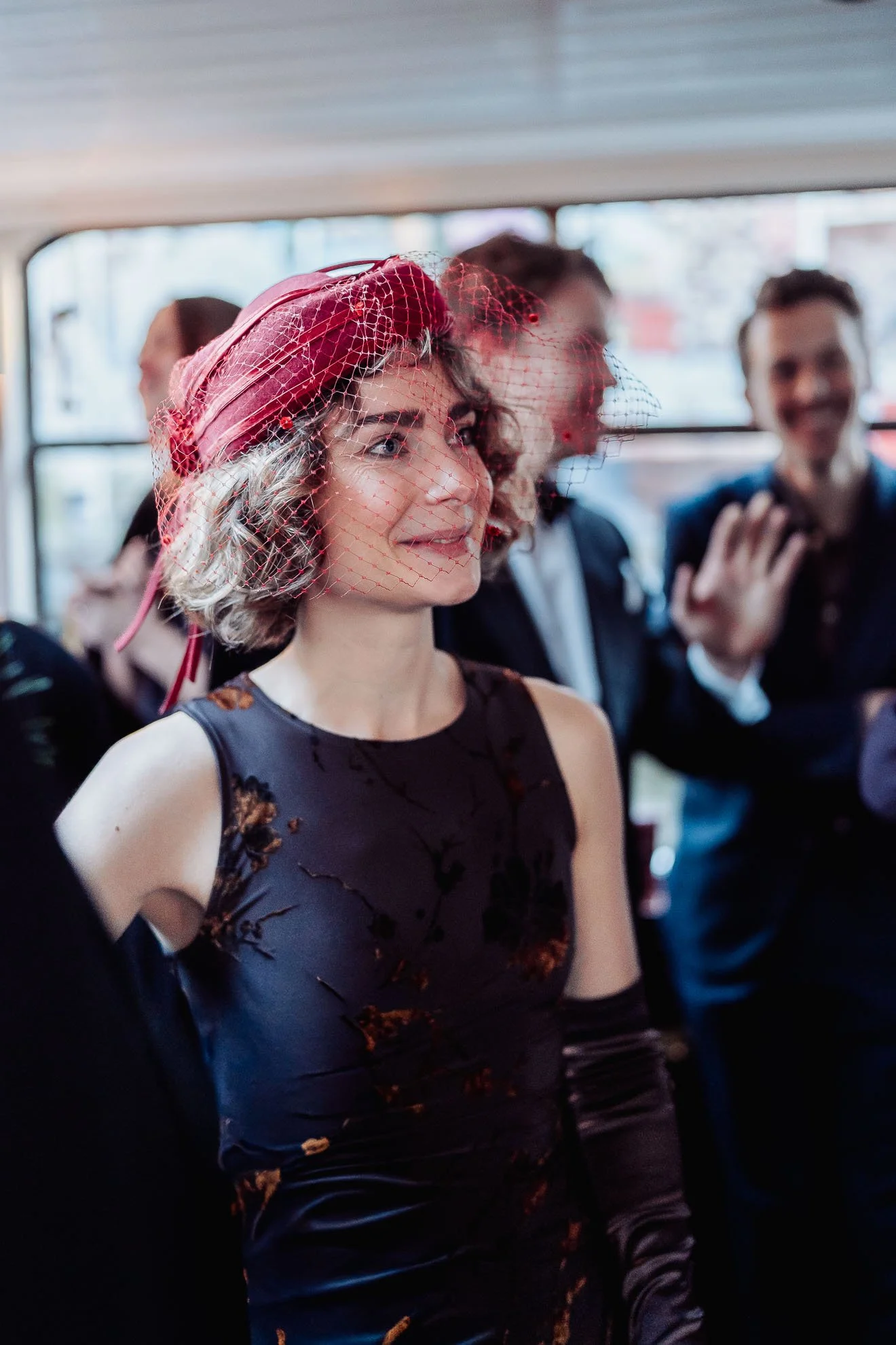 A woman in a floral dress with a red fascinator and birdcage veil, smiling on a bus. Other people are visible in the background, talking and smiling.