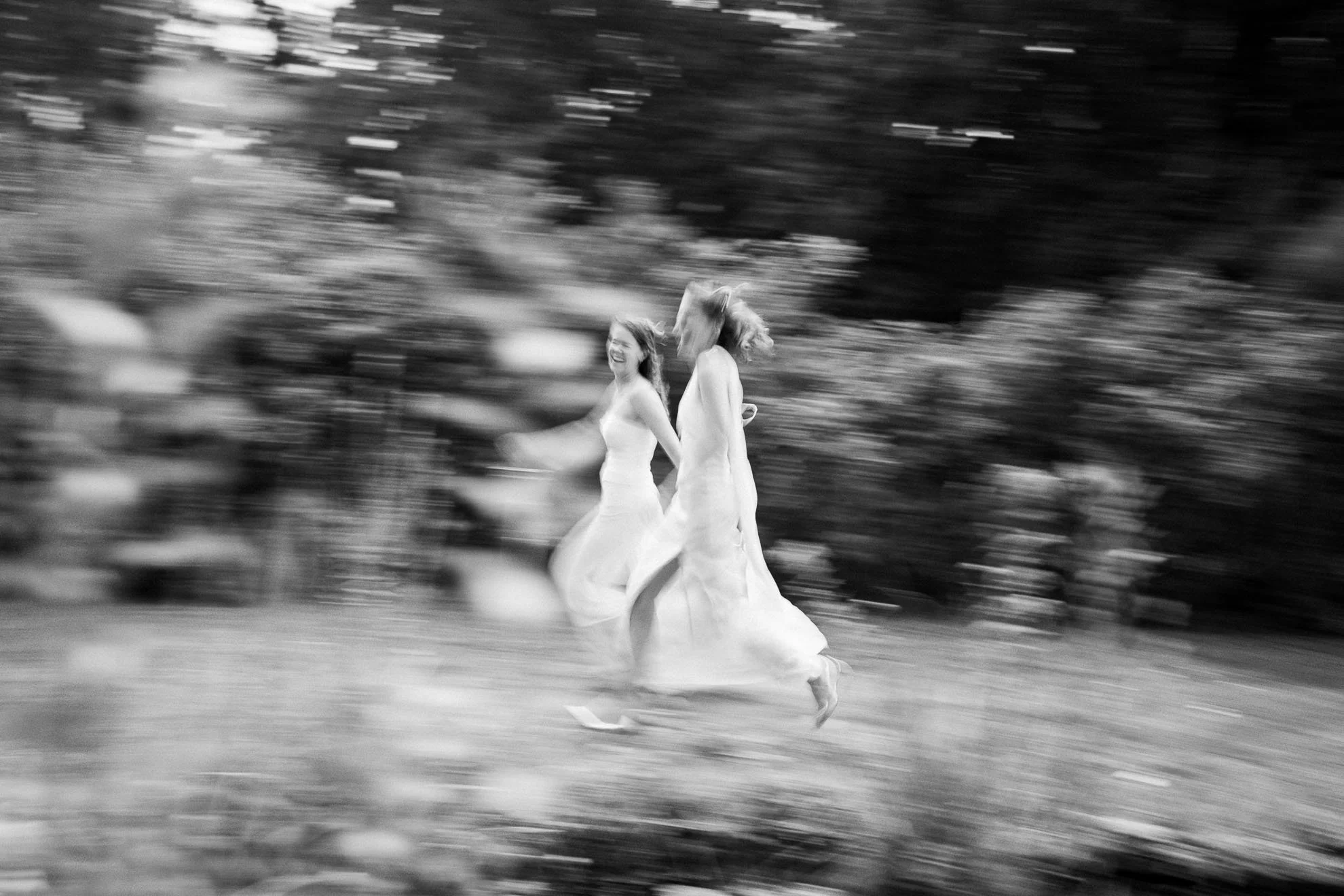 Black-and-white photo of two women in flowing dresses running outdoors, with trees and foliage in the background. The motion blur suggests they are moving quickly.