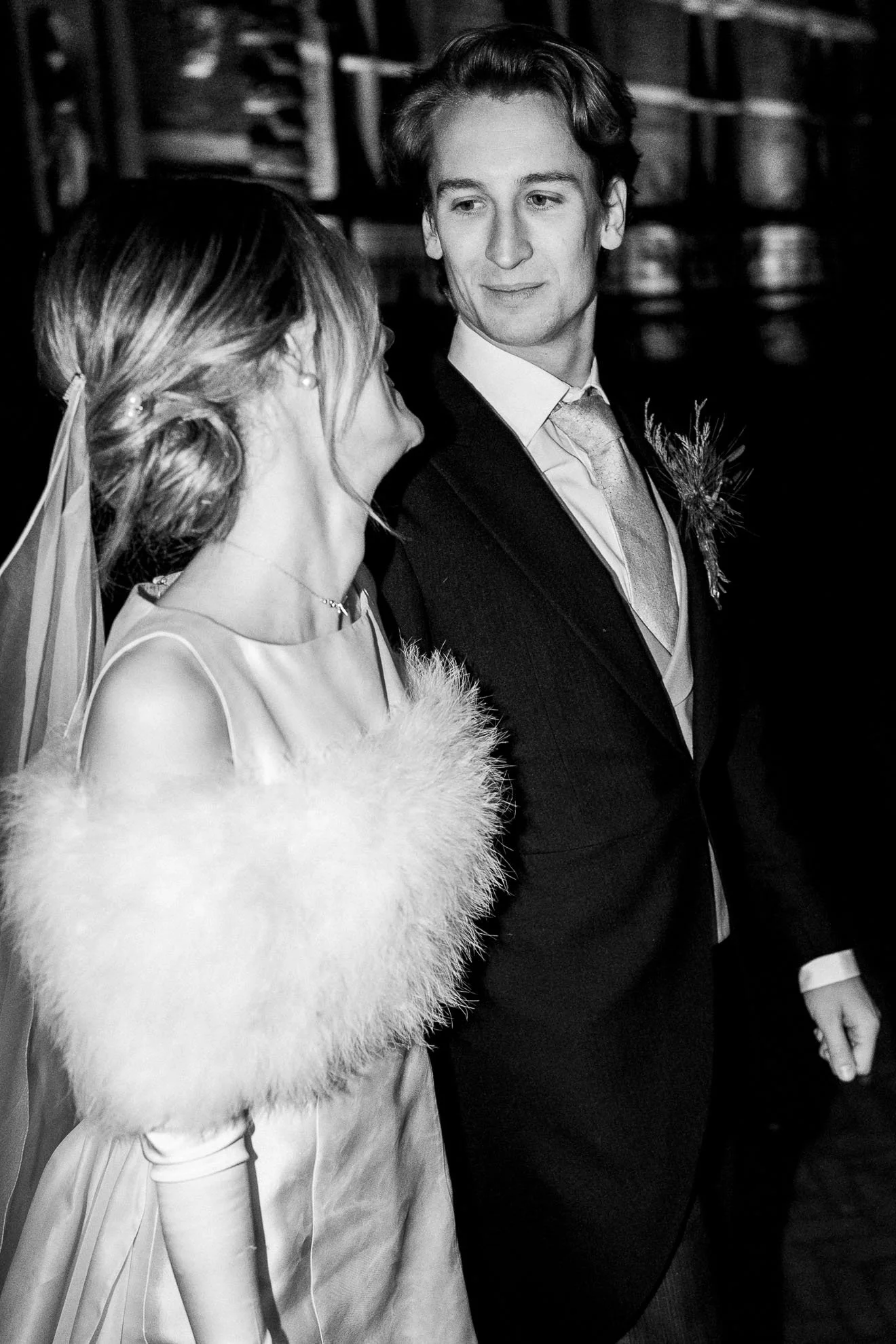 A black and white photo of a man and woman at a formal event, looking at each other. The woman is wearing a sleeveless dress with a fur wrap, and the man is in a suit with a tie and boutonniere.