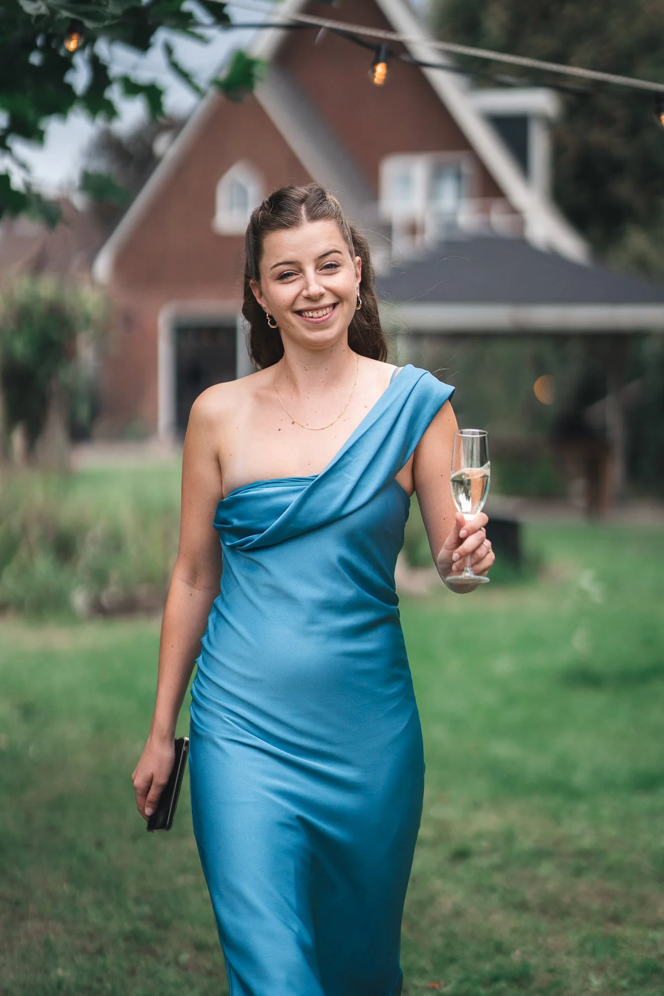 A woman in an elegant blue dress holds a champagne flute and smiles at an outdoor social event.