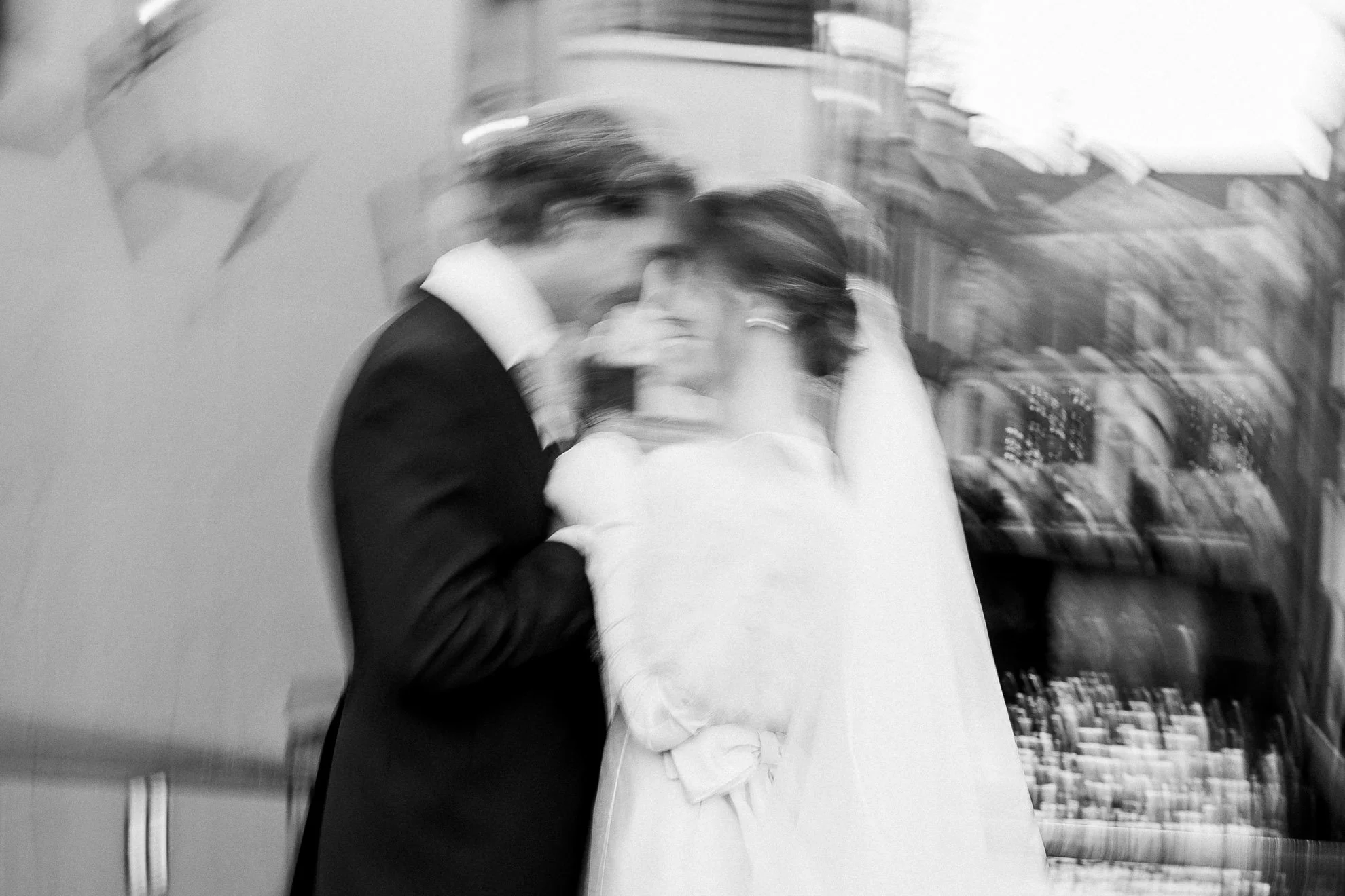 A blurry black and white photo of a man and a woman embracing and kissing. The man is wearing a suit and the woman is dressed in a light-colored outfit, possibly wedding attire. The background shows some books and shelves.