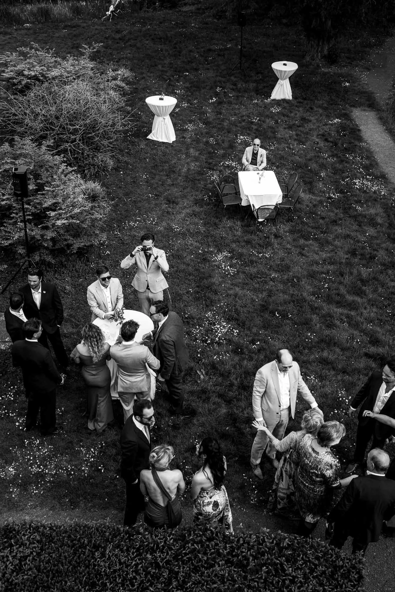 A black-and-white photo of a social gathering outdoors, with a group of people in formal attire standing and talking on a grassy area surrounded by bushes and trees, and two high-top tables covered with tablecloths in the background.