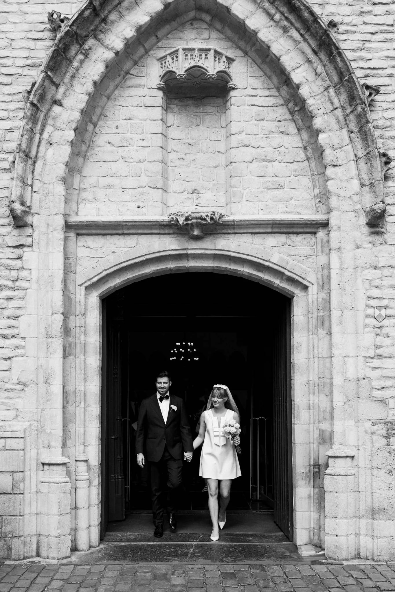 Een zwart-wit foto van een pasgetrouwd stel dat door de ingang van een oude kerk loopt. De man draagt een zwart pak met een witte strohoed, de vrouw draagt een witte jurk met een bloemenboeket.