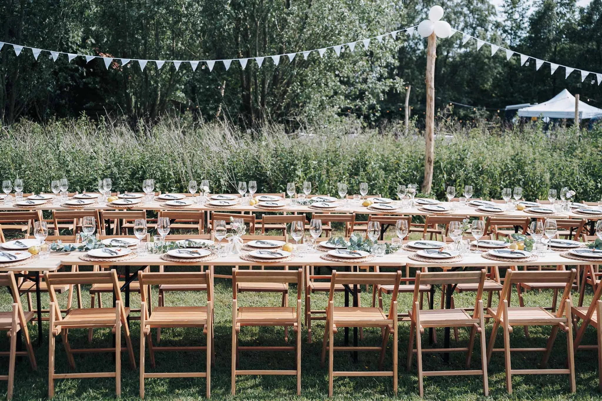 Een feestopstelling met lange houten tafels en stoelen, versierd met glazen, borden, bestek en decoratie, buiten in een groene tuin met bomen, vlaggenlint en een tent op de achtergrond.