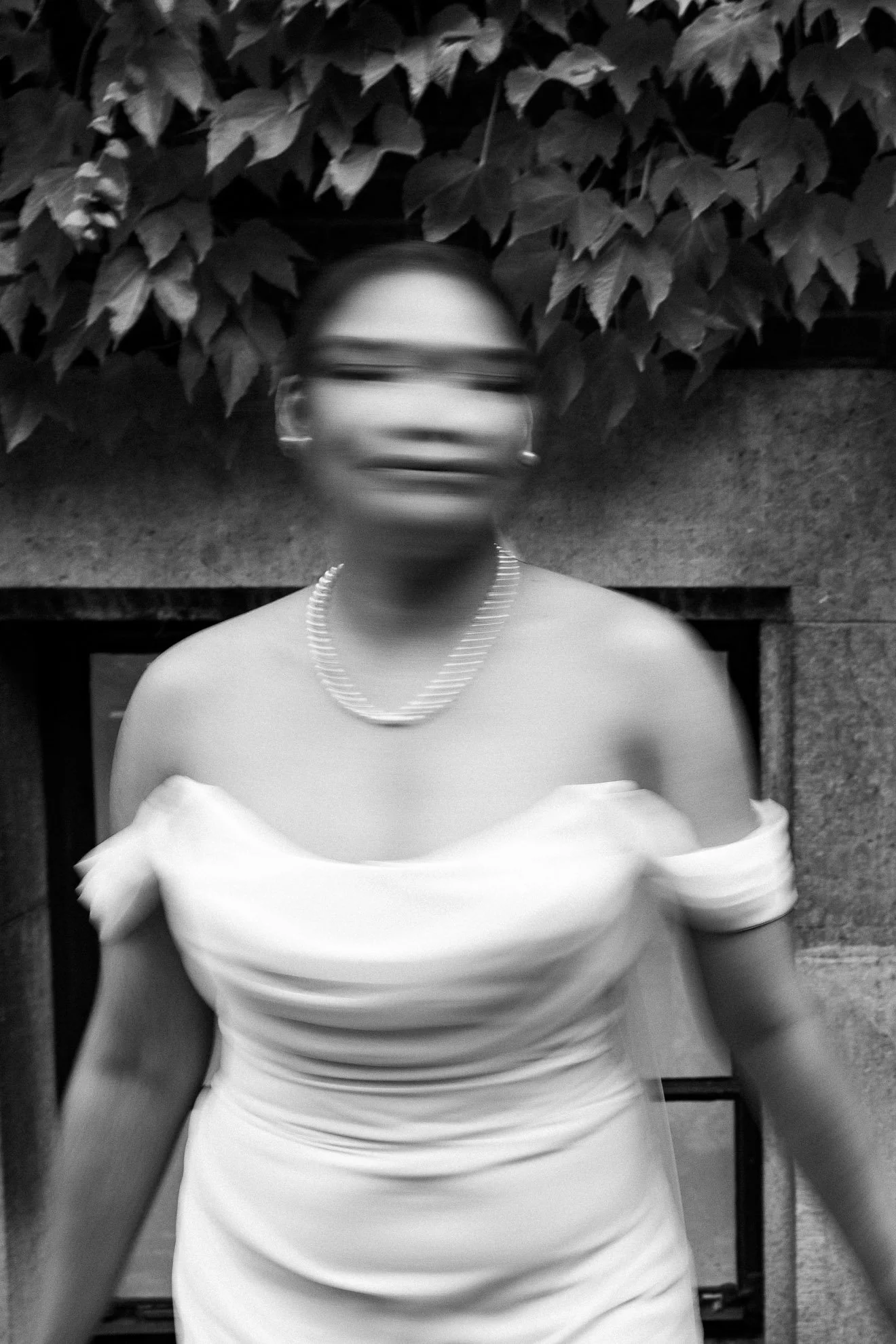 A person wearing glasses and a pearl necklace, dressed in an off-shoulder white dress, standing in front of a leafy background, with motion blur creating a sense of movement.