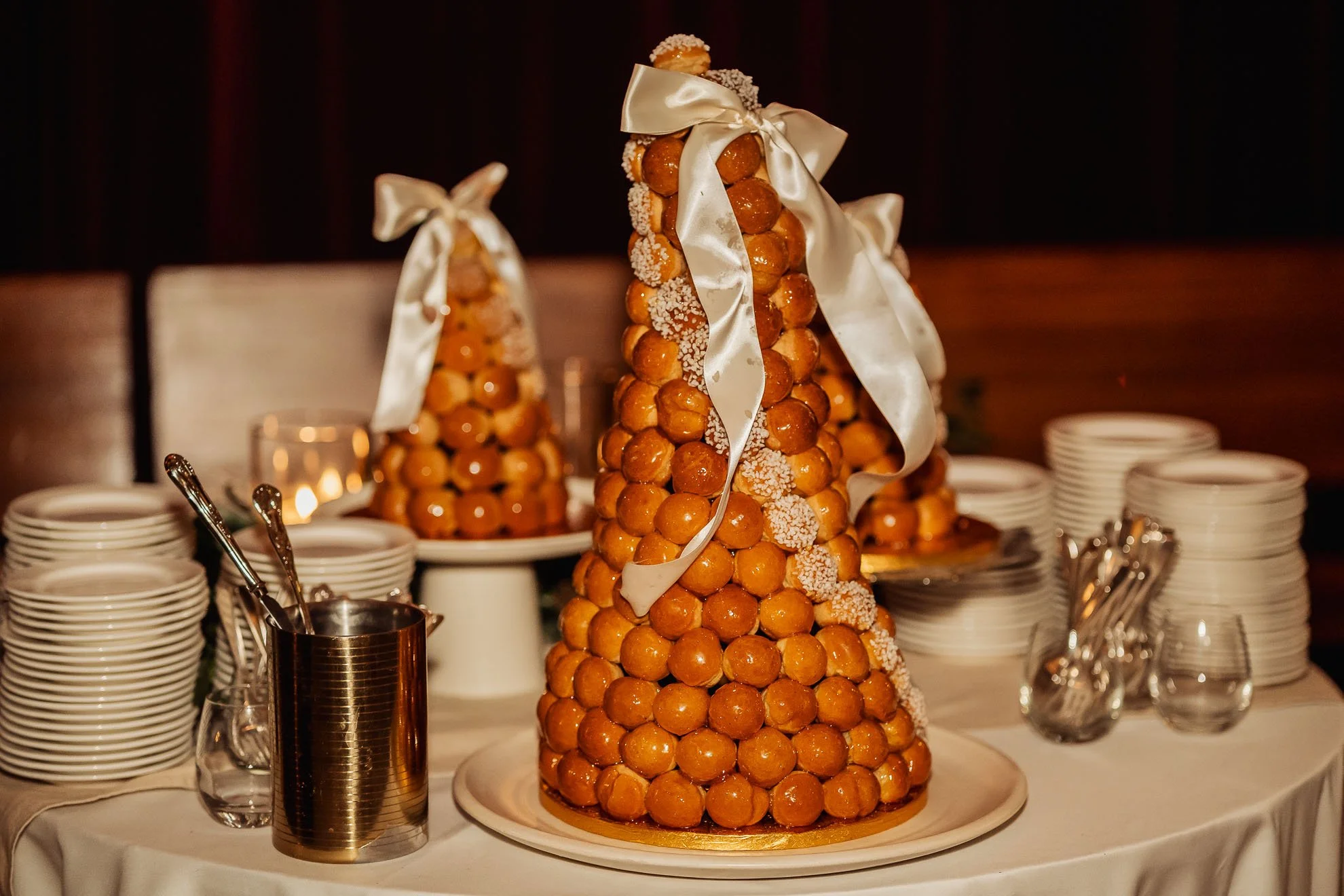 A croquembouche decorated with white ribbons and topped with profiteroles, displayed on a table with stacks of white plates and bakery utensils.