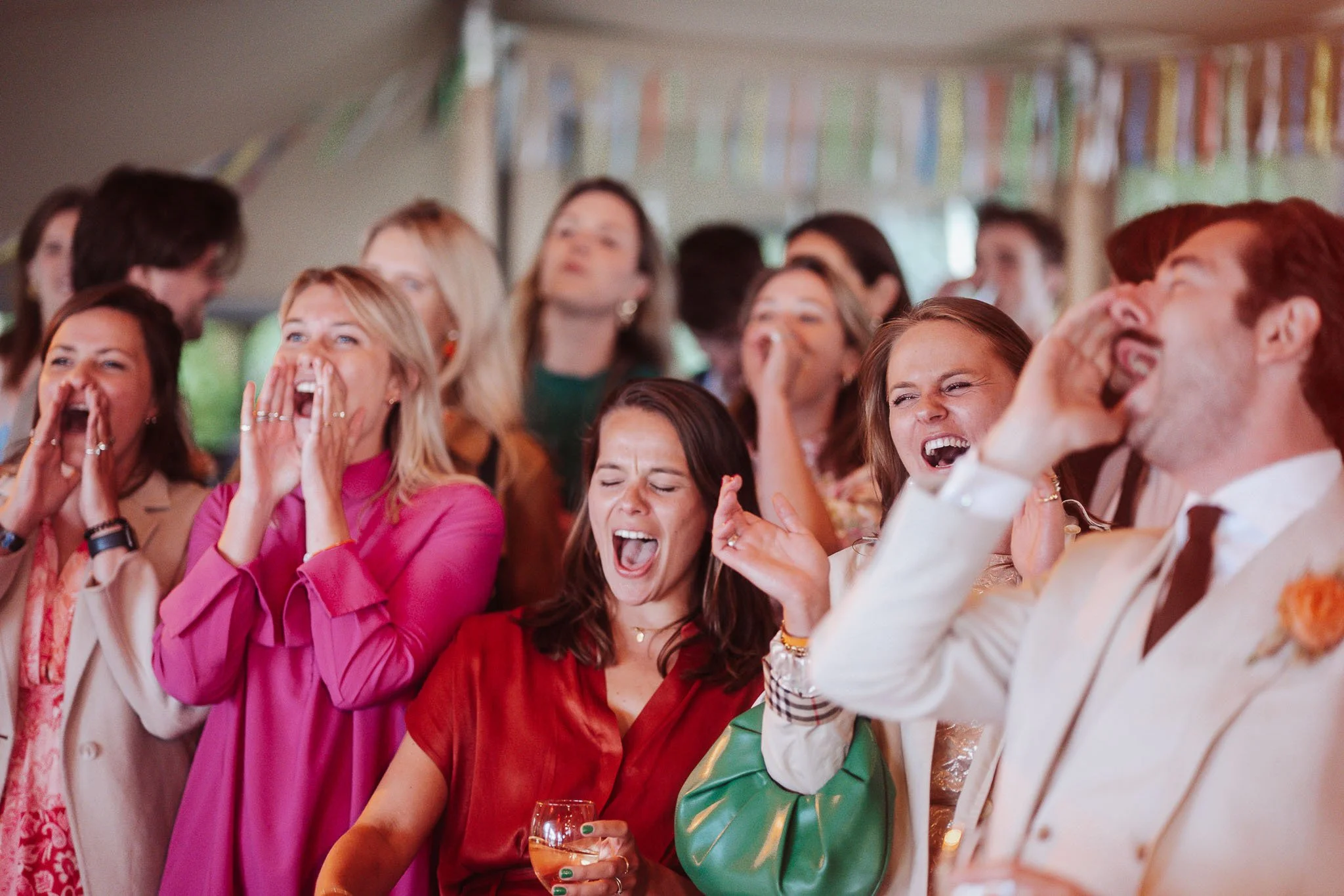 Een groep mensen die lachen en plezier maken op een feest of bruiloft, onder een tent met kleurrijke vlaggetjes.