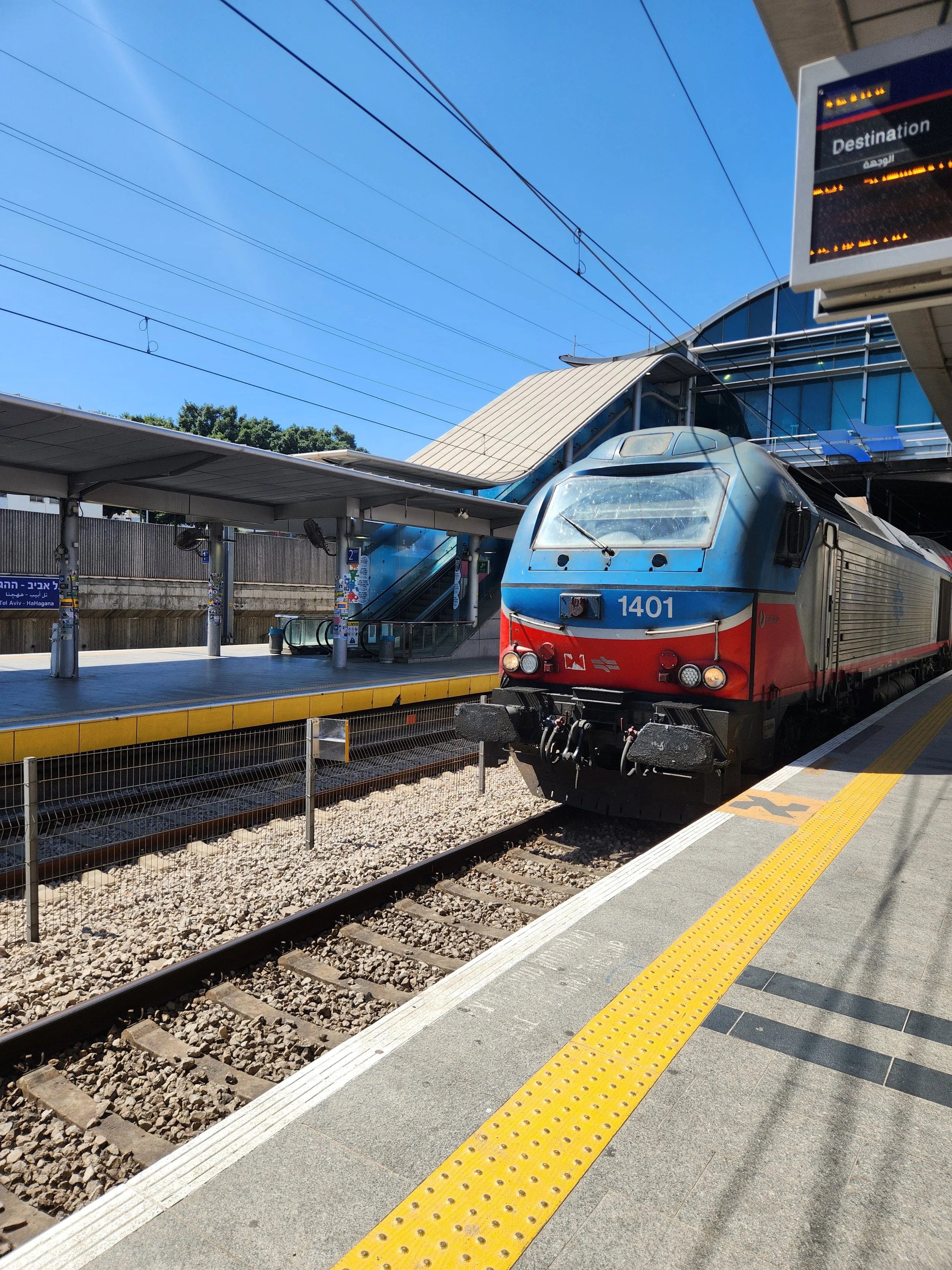Passenger train at a modern train station platform under a blue sky, with overhead wiring overhead, a yellow tactile strip along the edge of the platform, and an electronic schedule display.